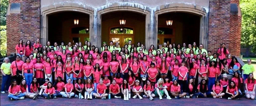 Group of young women and a few staff members standing and sitting in front of a brick building, all wearing matching pink and neon green t-shirts.