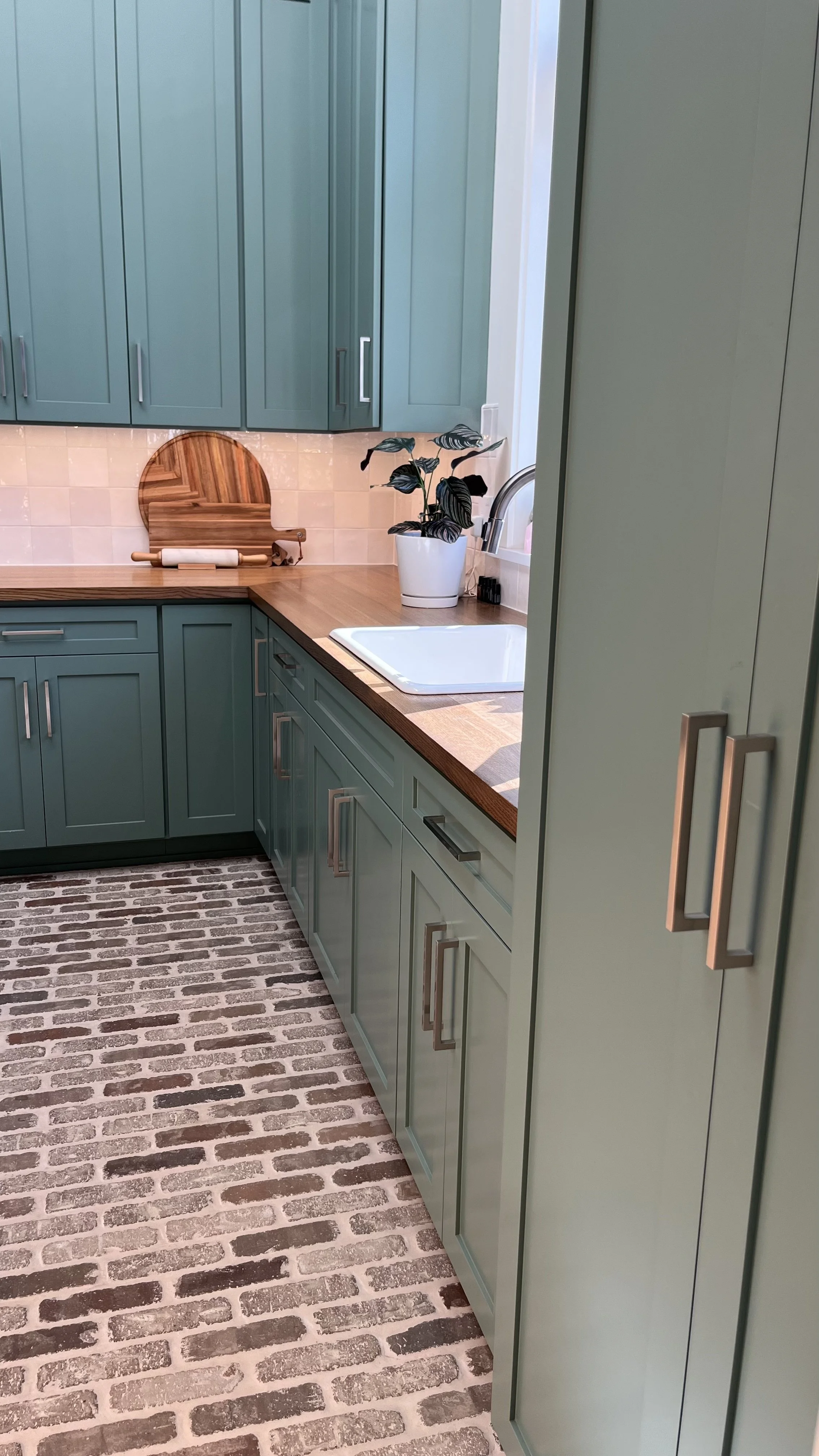 A kitchen with green cabinets, a wooden countertop, a white sink, a potted plant, a wooden cutting board with a rolling pin, and brick-patterned flooring.