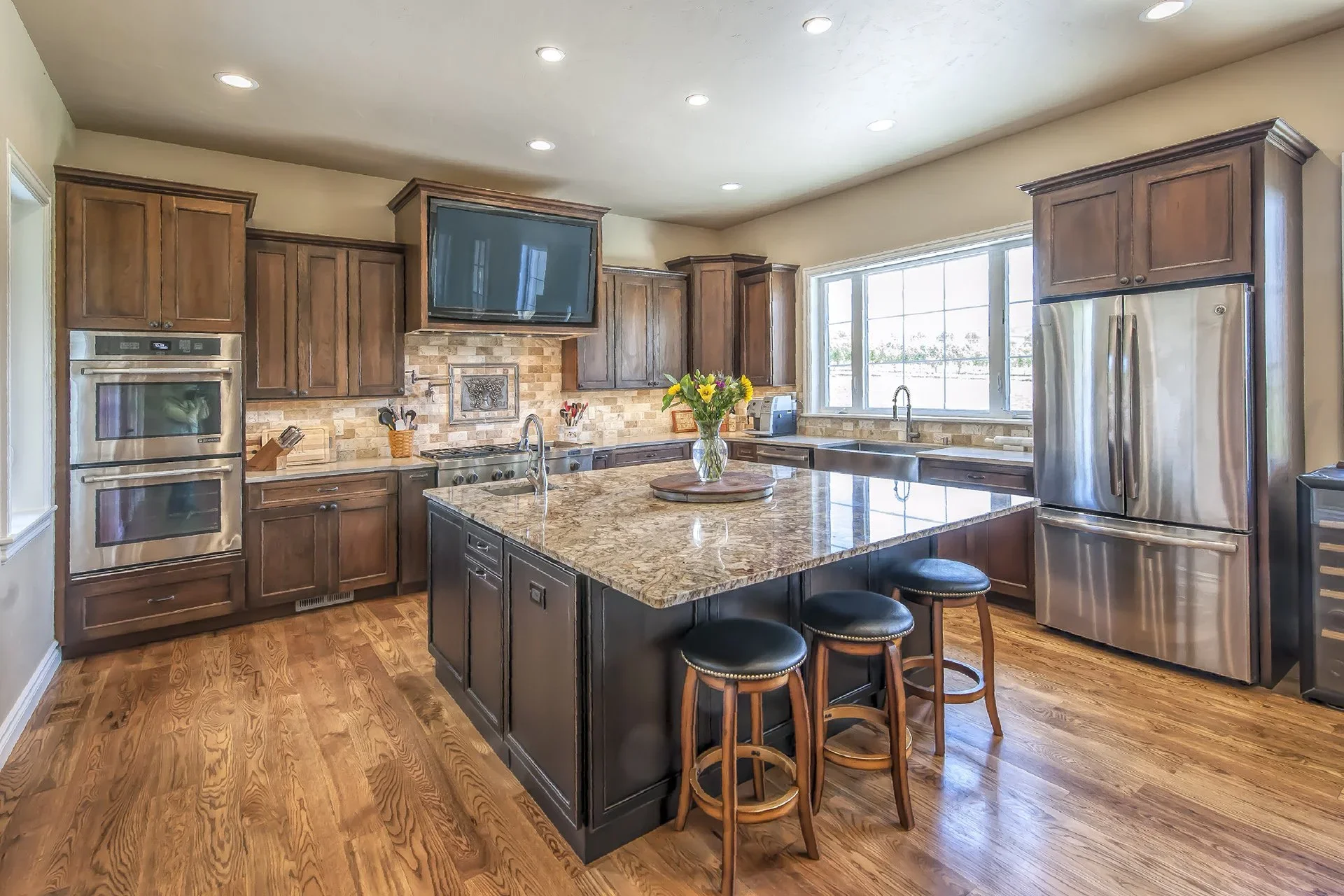 Modern kitchen with wooden cabinets, granite island with bar stools, stainless steel appliances, large window, and hardwood flooring.