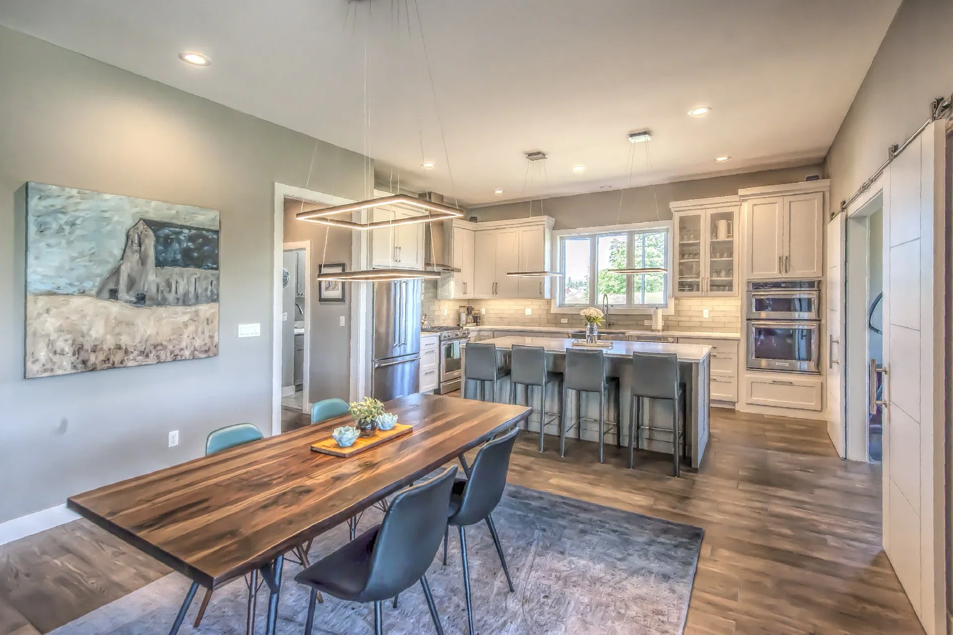 Open-concept kitchen and dining area with a wooden dining table, teal chairs, and contemporary kitchen cabinetry, appliances, and a window overlooking trees.