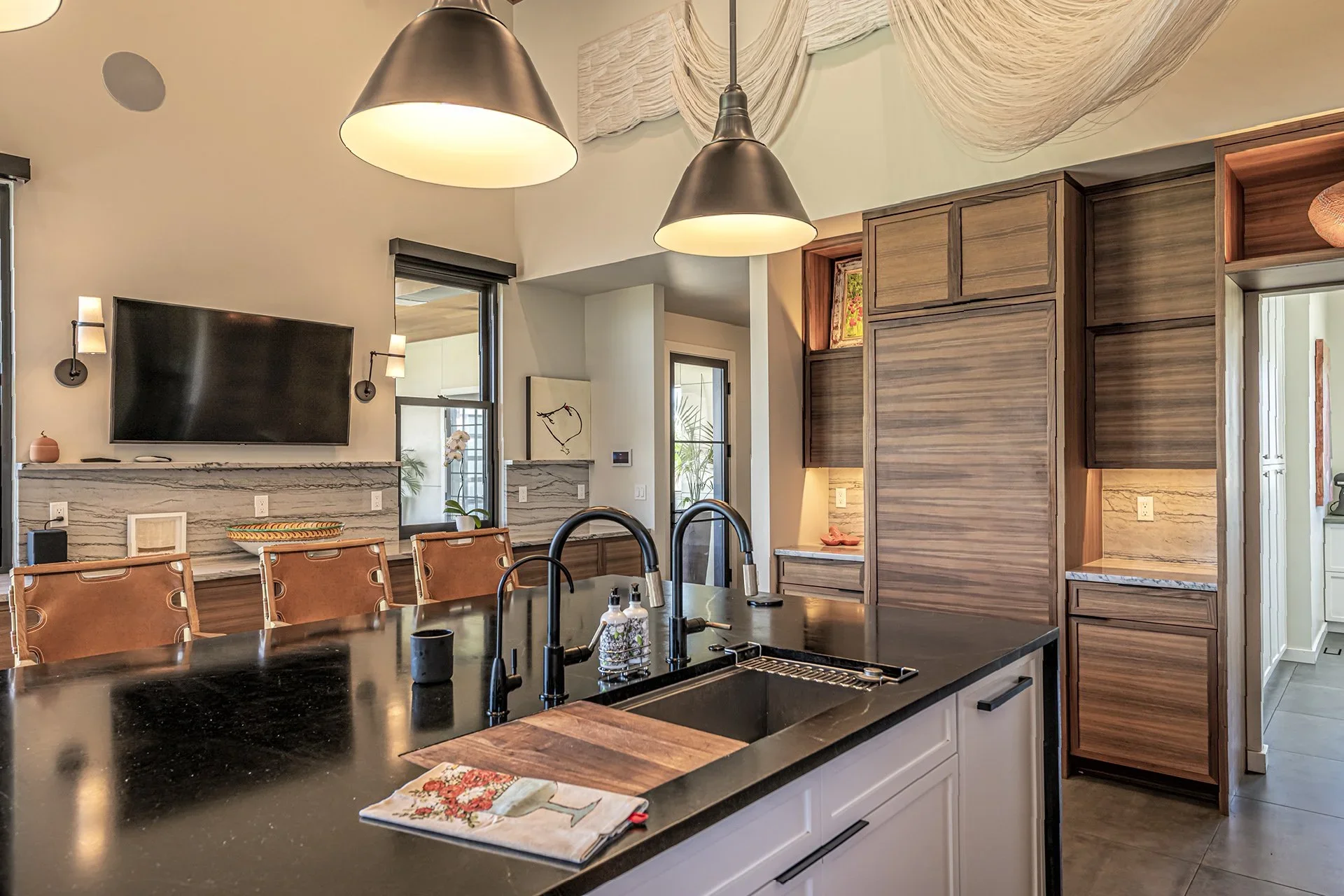 Modern kitchen with black countertop, wooden cabinets, and two pendant lights, adjacent to a living space with a mounted TV and art on the wall.