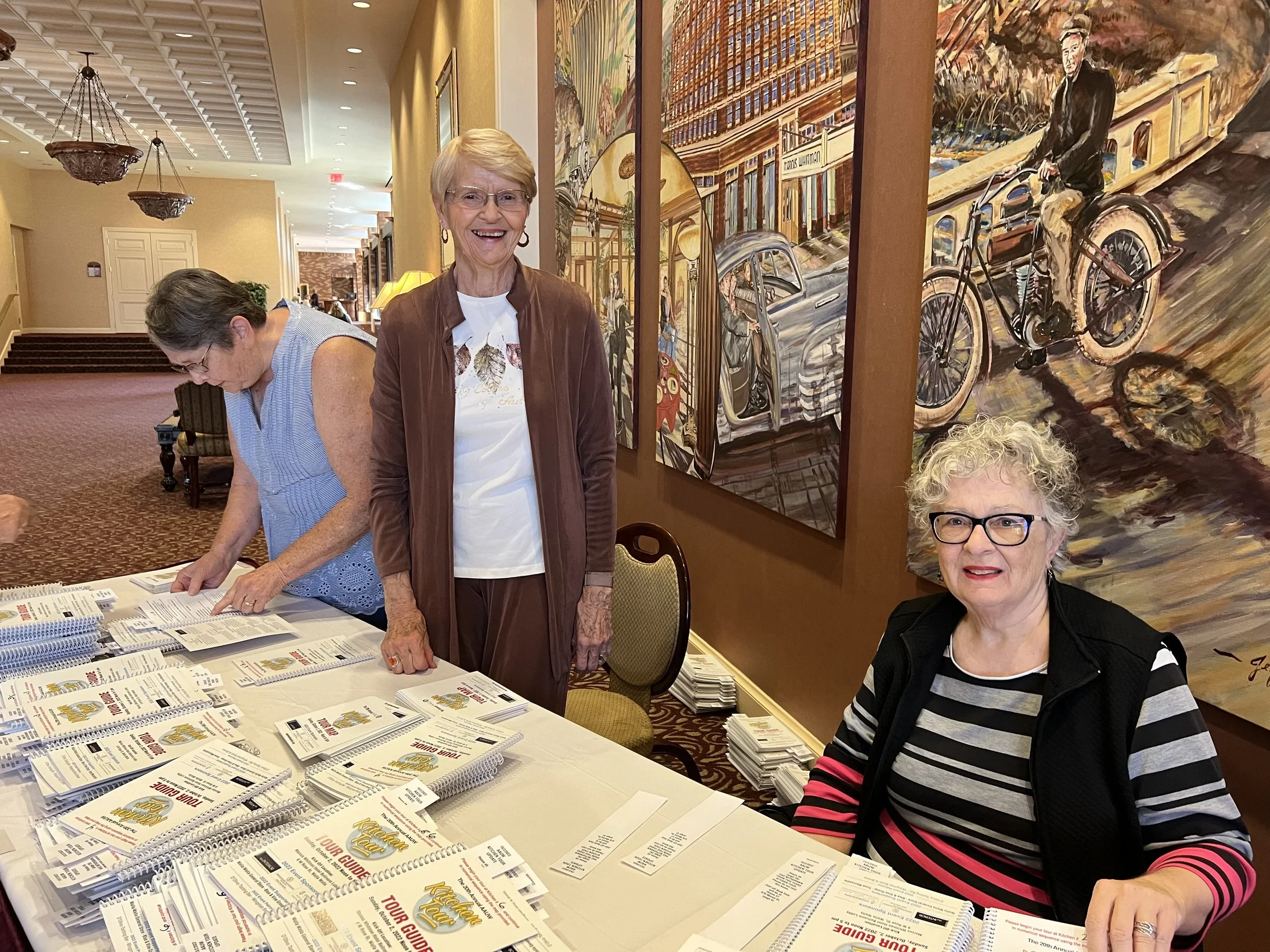 Three older women in a hotel lobby, with two of them standing and one sitting, surrounded by pamphlets and brochures on a table. The standing woman is smiling at the camera, wearing glasses and a brown jacket. The sitting woman has curly white hair, glasses, and is wearing a striped black, white, and pink top. There are large, colorful paintings on the wall behind them, and another woman in a sleeveless top is looking down at the table.