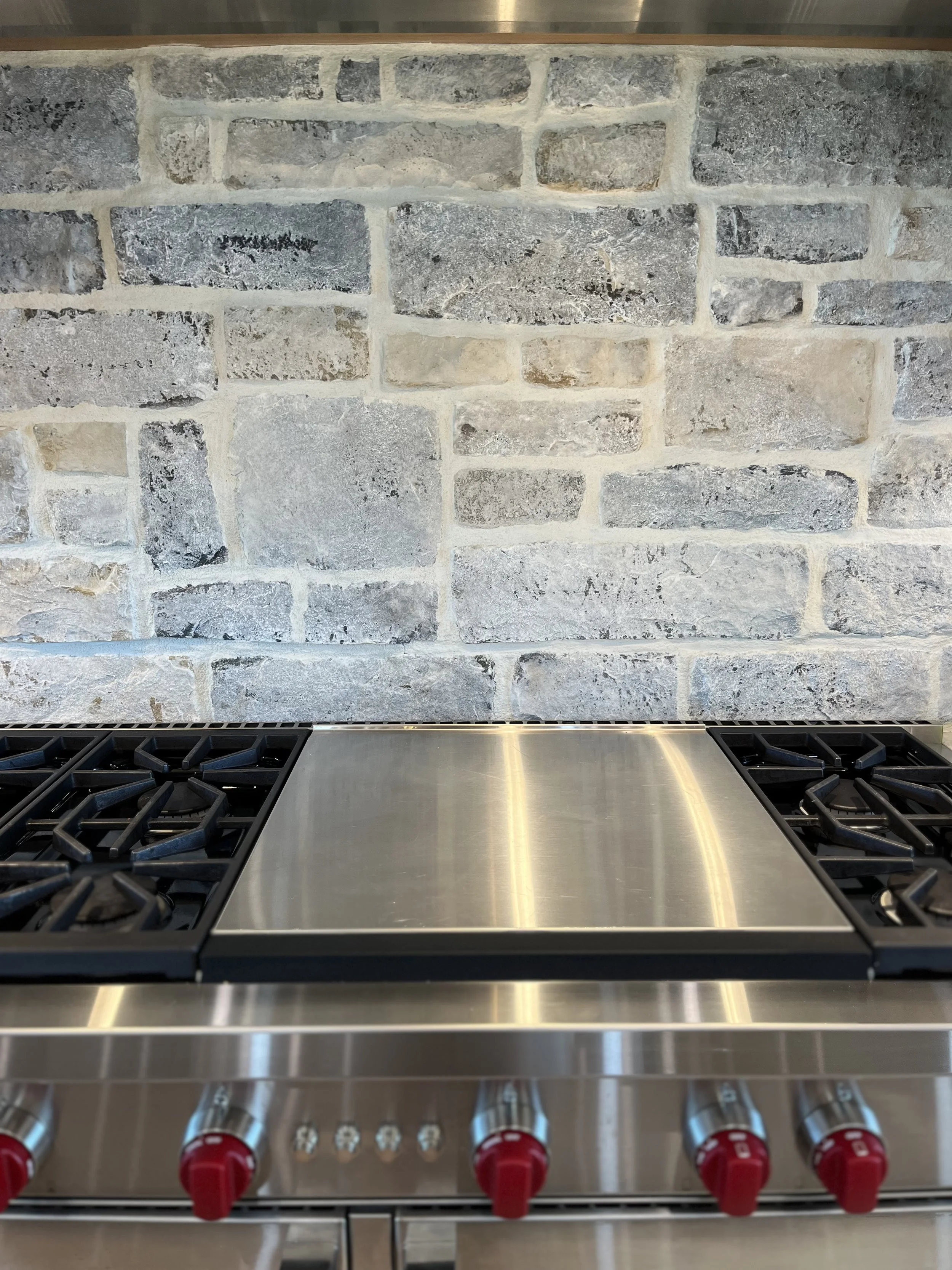 A modern stainless steel kitchen stove with four burners and red control knobs, set against a gray and black brick backsplash.