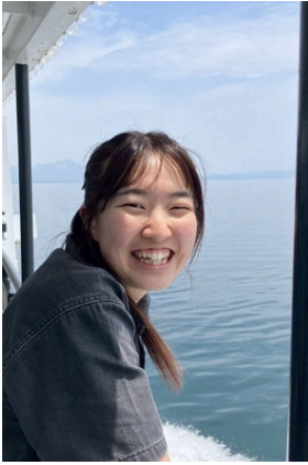 A young woman smiling with water and a blue sky in the background, on a boat or near a water body.