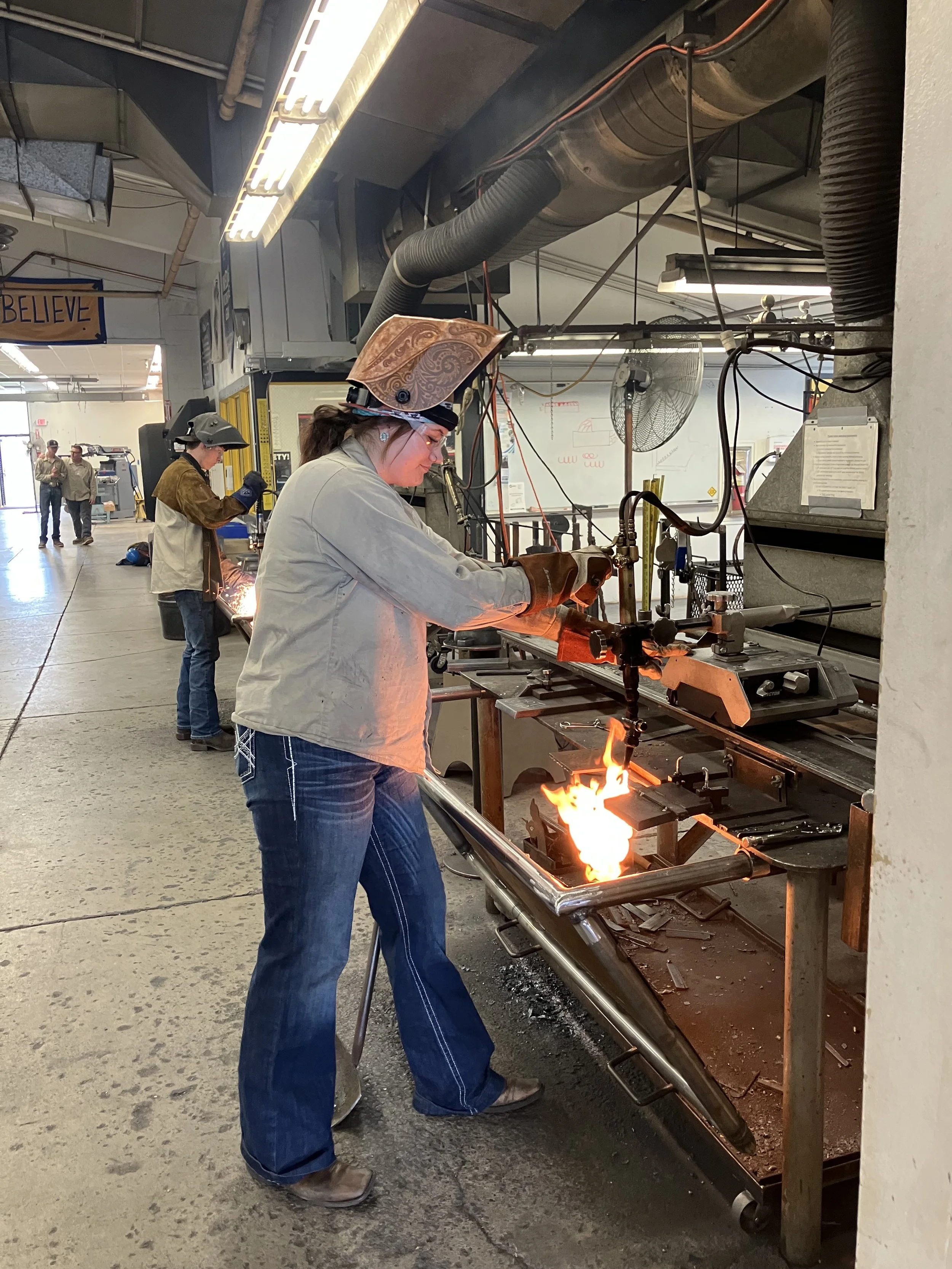 Woman working with welding tools on a metal pipe or rod in an industrial workshop, wearing protective glasses, a welding helmet pushed back, gloves, and casual work clothes. Sparks and flames are visible from the welding process, with another worker in the background also engaged in similar work.