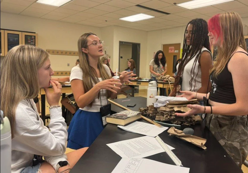 Four young women are engaged in a discussion in a classroom or lab. One is standing and talking, holding papers; the others are listening, with one seated and two standing. There are papers, books, and supplies on the table.