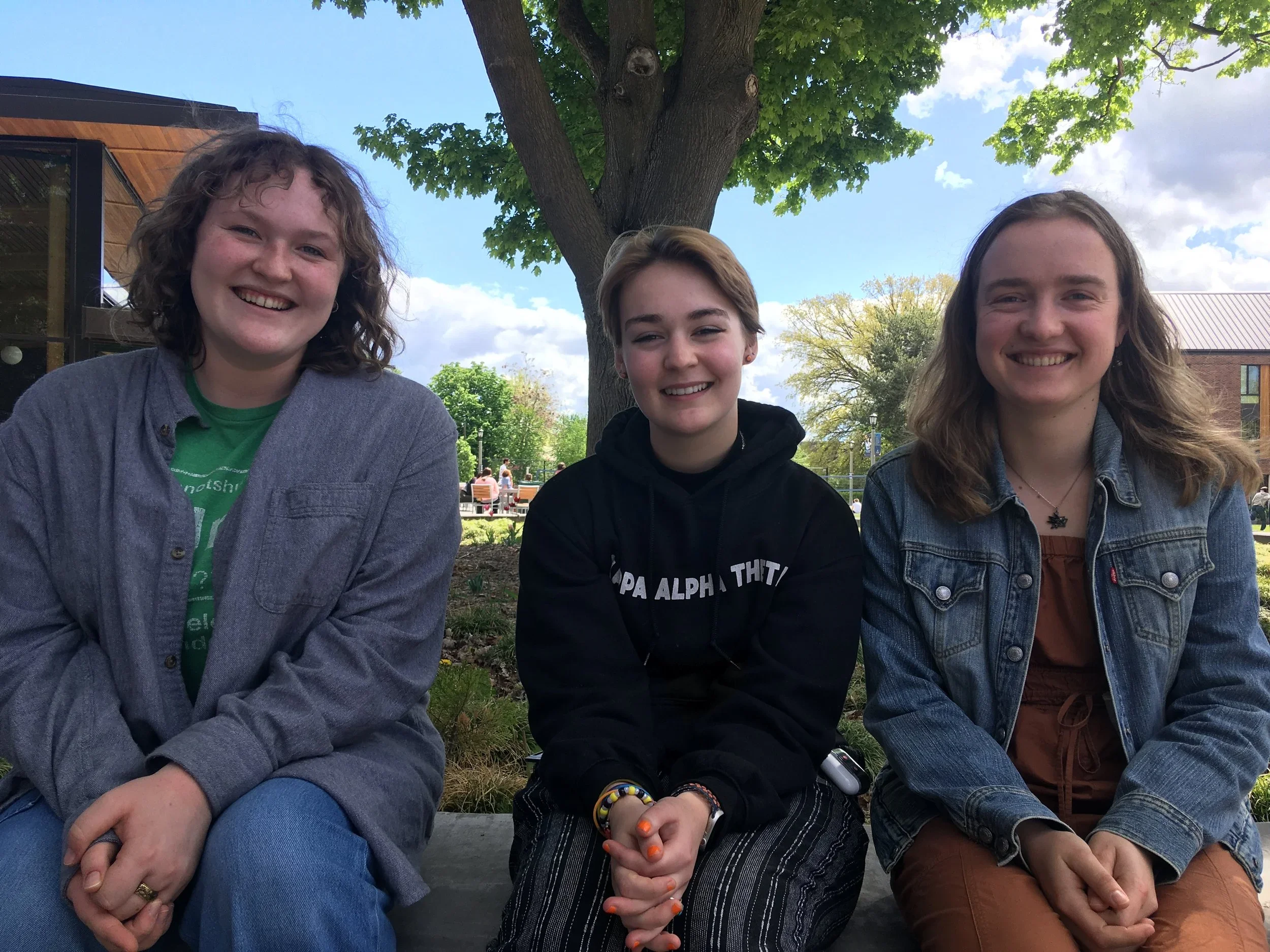 Three young women sitting outside in front of a large leafy tree. They are smiling and enjoying the sunny day, with buildings and other people in the background.