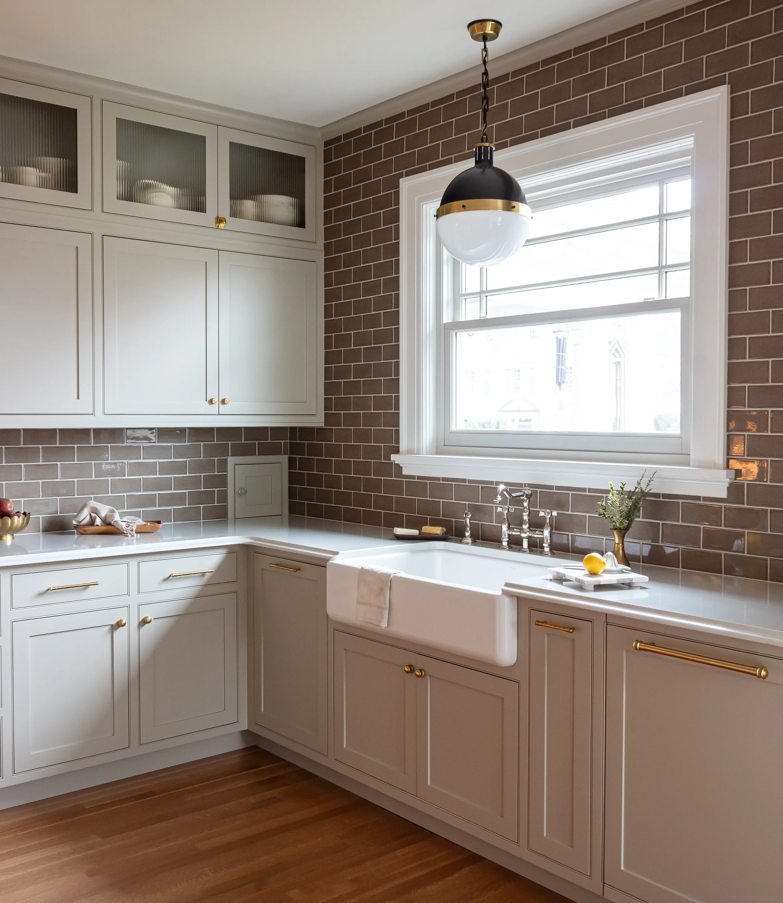 A kitchen with white cabinets, a large window, a black and gold hanging light fixture, a white farmhouse sink, a small potted plant, lemons, and a brick wall.