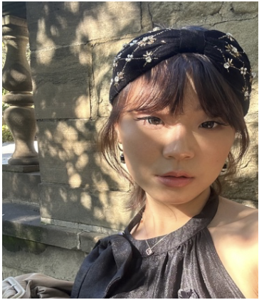 Woman wearing a black headband with floral embroidery, standing outdoors against a stone wall with shadows.