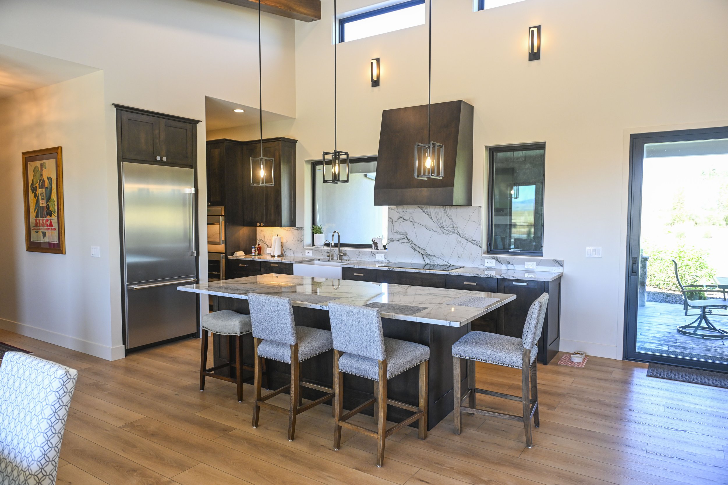 Modern kitchen with dark wood cabinets, white marble countertops, and stainless steel appliances. There is a kitchen island with seating and pendant lighting, large windows, and a sliding glass door leading to an outdoor deck.