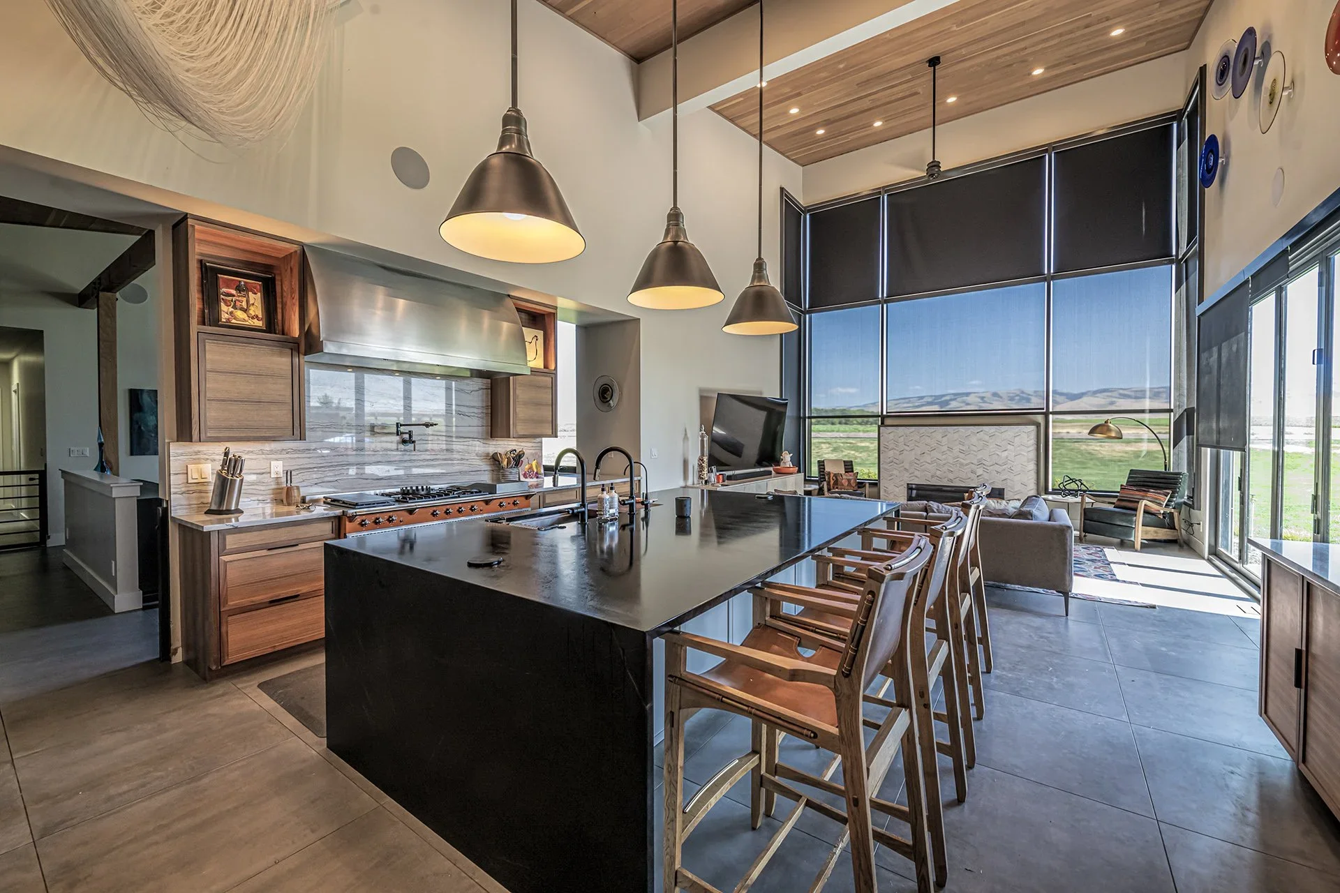 Modern open-concept kitchen and living area with large windows, wooden ceiling, pendant lights, kitchen island, and seating area with armchairs and a TV.