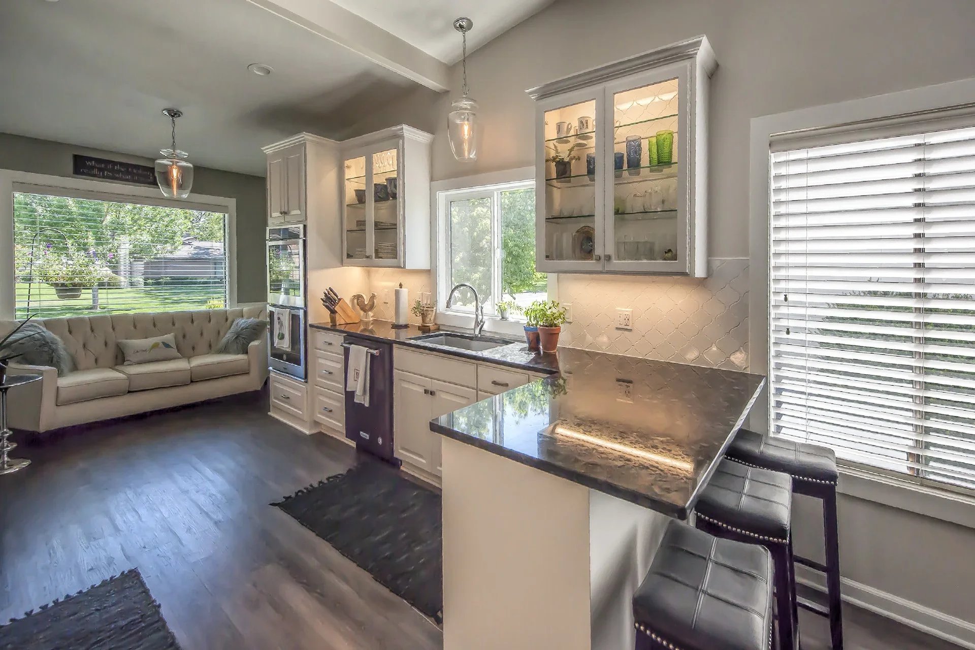 A bright kitchen with white cabinets, granite countertops, and a window above the sink. There are glass-front cabinets displaying dishes and cups, with a black granite island counter. To the left, a beige tufted sofa with pillows is in front of a lar