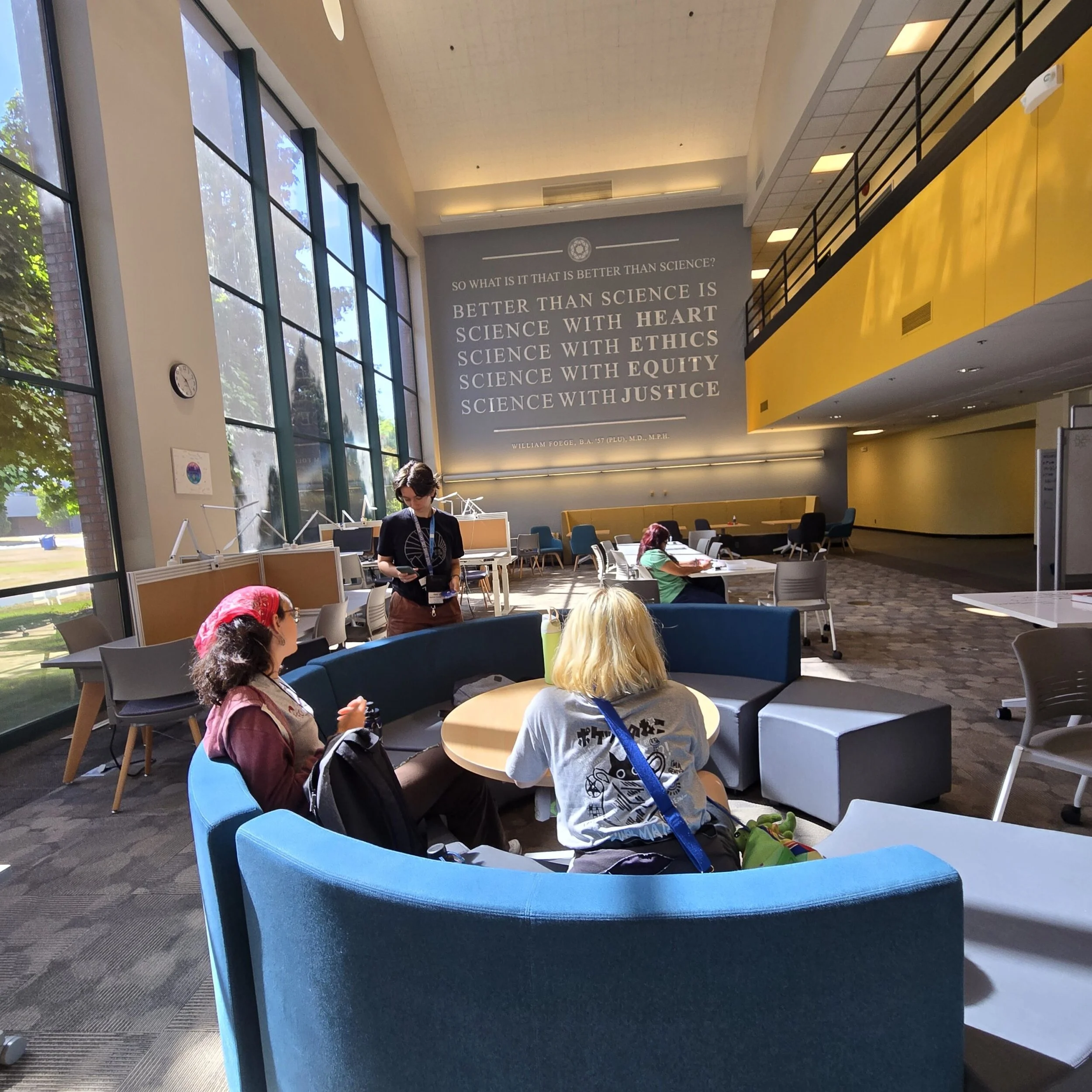 The image shows the interior of a modern building with high ceilings, large windows, and open space. Several people are sitting or standing, engaged in activities. A large gray wall displays a quote about science and ethics. The building has a yellow
