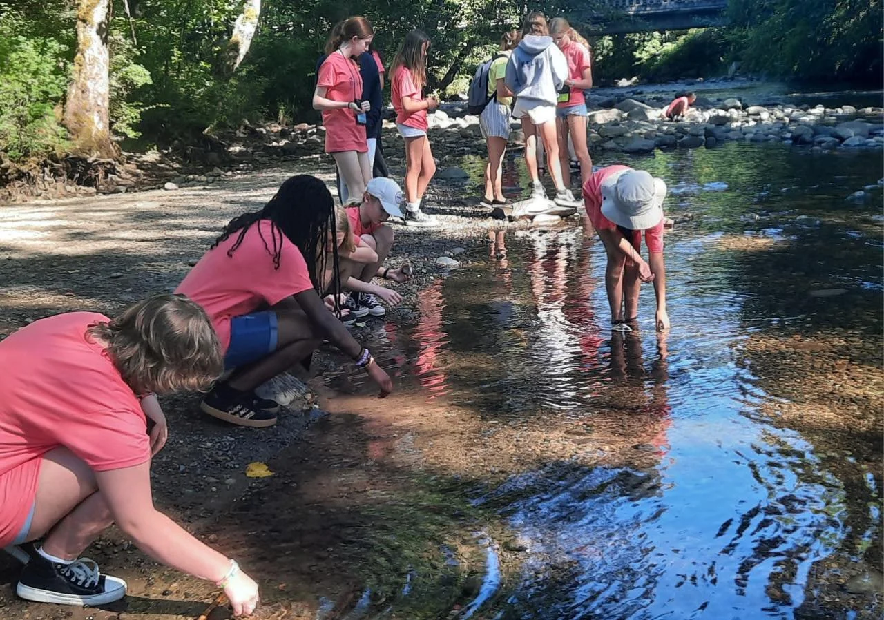 A group of children and teenagers gather by the edge of a river, some crouching and reaching into the water, while others stand on the shoreline in a wooded area under sunlight.