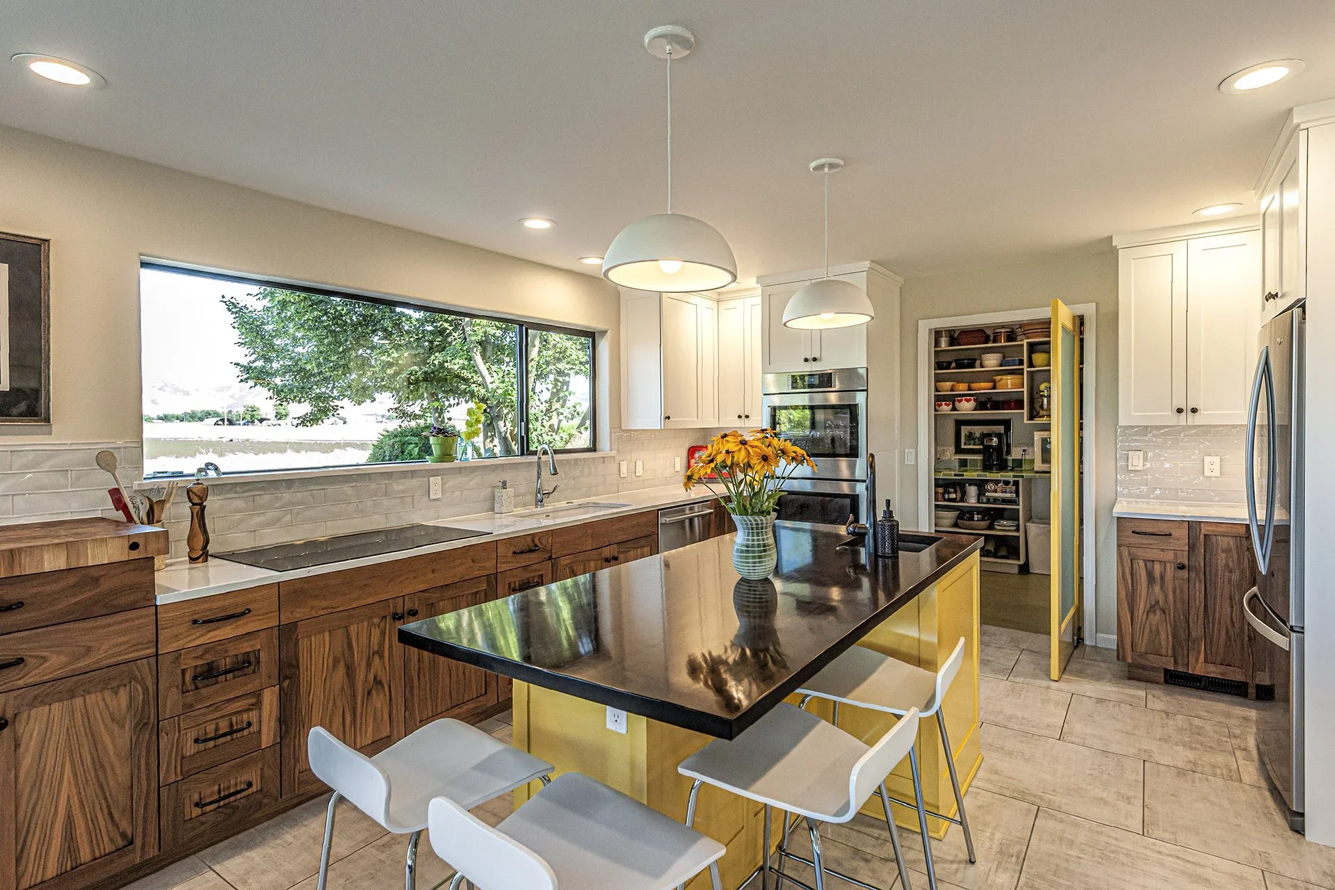 Modern kitchen with white cabinets, wooden drawers, black kitchen island with yellow base, large window showing greenery outside, stainless steel appliances, and a black countertop with a vase of yellow flowers.