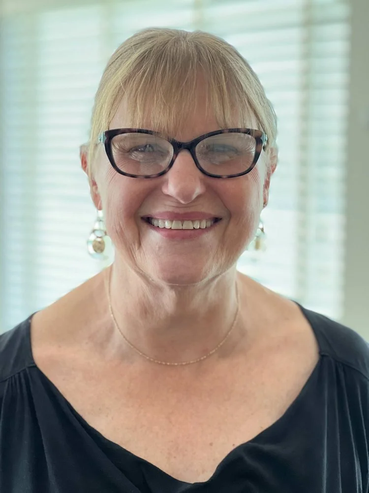 A smiling middle-aged woman with blonde hair, wearing glasses, earrings, and a black top.