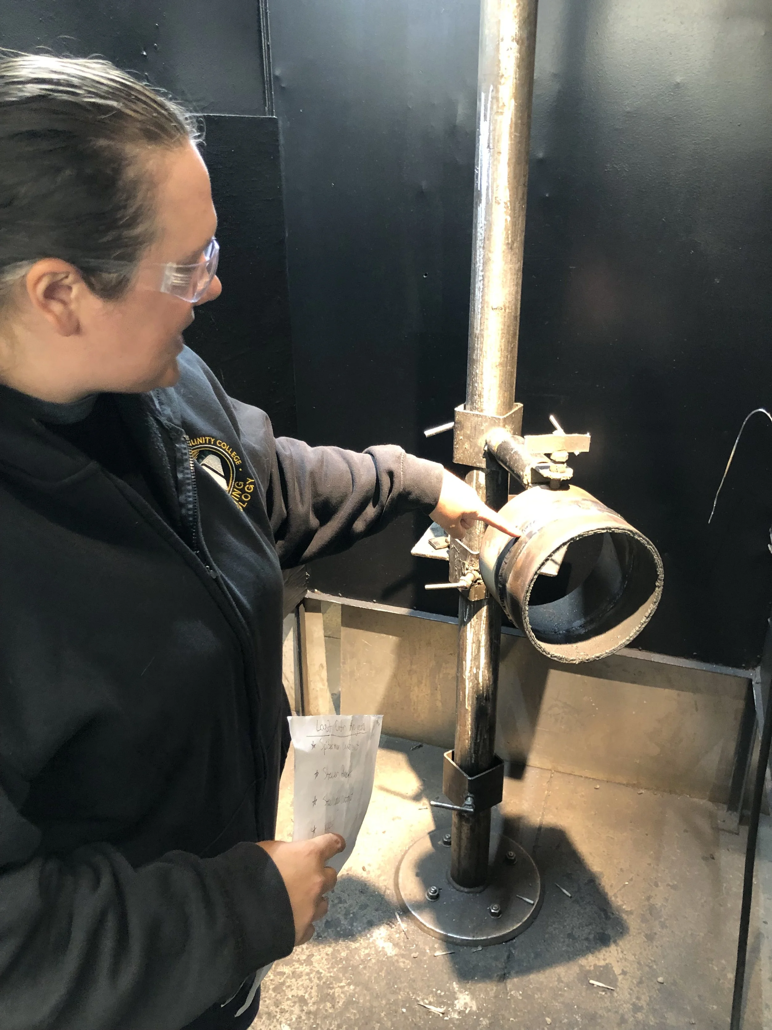 A woman wearing safety glasses and a jacket with a patch on it is working on a metal structure in an industrial setting.
