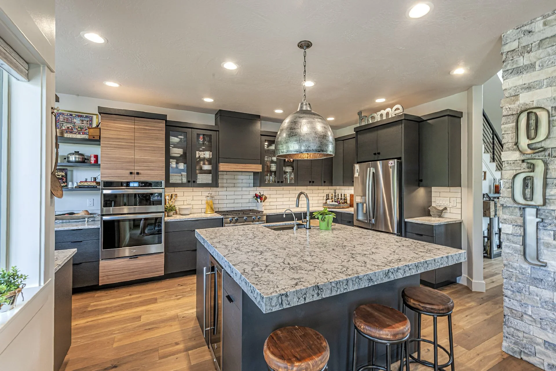 Modern kitchen with black cabinets, white marble countertops, stainless steel appliances, a central island with bar stools, and a brick wall accent. Overhead pendant light and recessed lighting.
