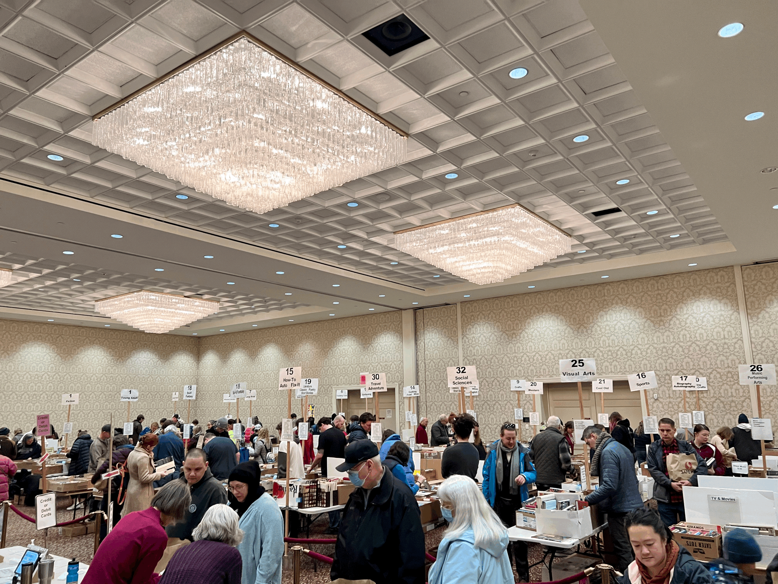 A large indoor event with many people browsing tables filled with books and other items, under decorative tile ceiling with large rectangular chandeliers.
