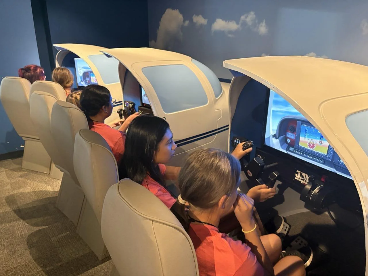 Children sitting in flight simulator cabins with cockpit controls, screens, and overhead canopies, simulating airplane flying experience.