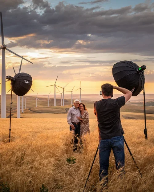 A man takes a photograph of an elderly couple in a wheat field at sunset, with wind turbines in the background.