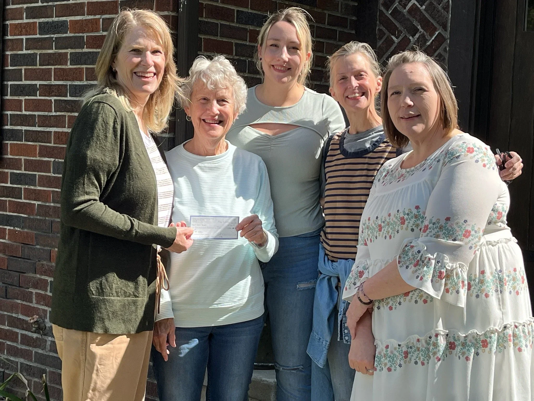 A group of six women standing outside in front of a brick wall, smiling, with one woman holding a check.