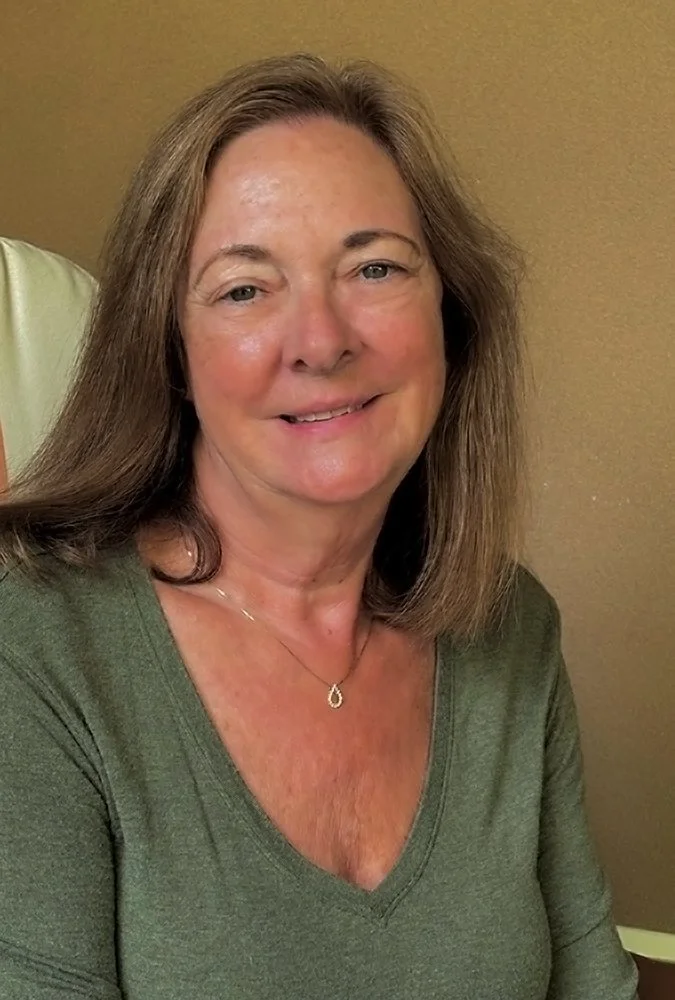 A woman with shoulder-length brown hair, wearing a green V-neck shirt and a delicate necklace, smiling at the camera against a neutral background.