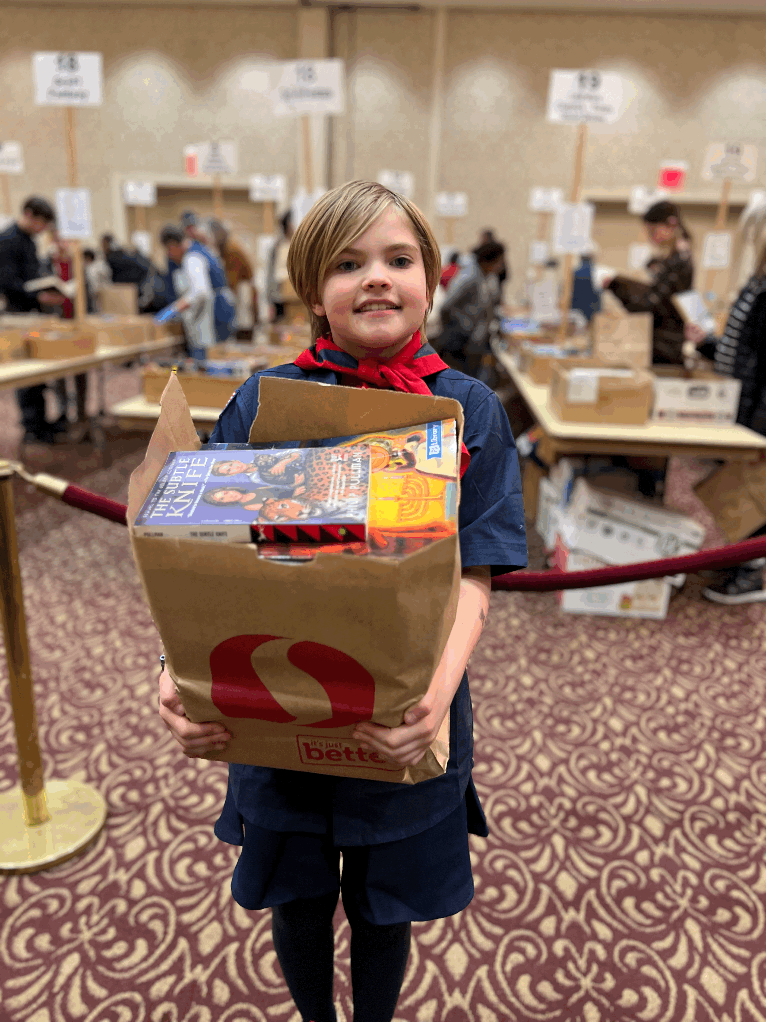 A young girl dressed in a scout uniform holding a shopping bag with books inside at an indoor bookstore or library.