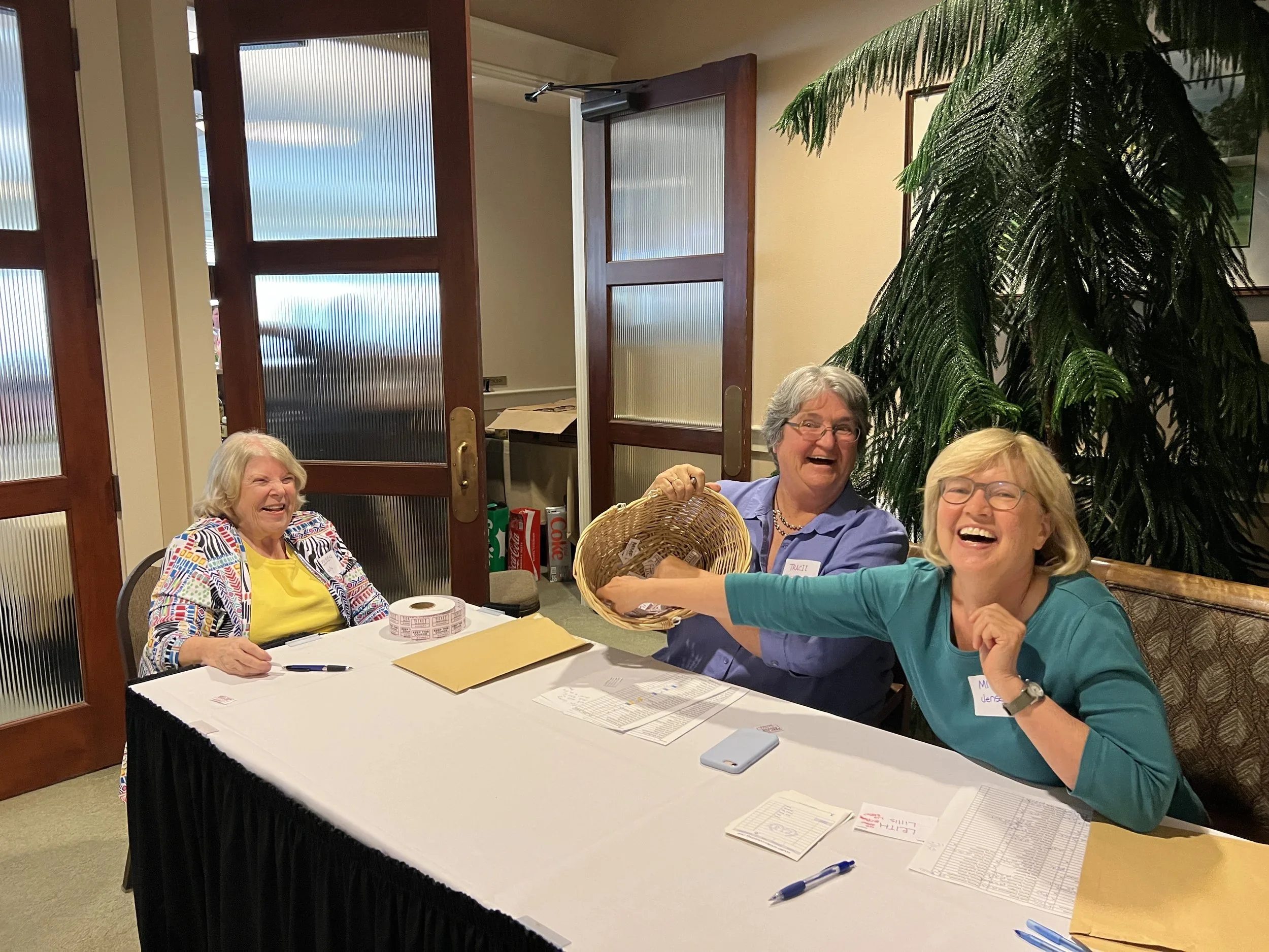 Four women sitting around a table inside a room, smiling and laughing, with papers and pens in front of them. There is a large green plant behind them.