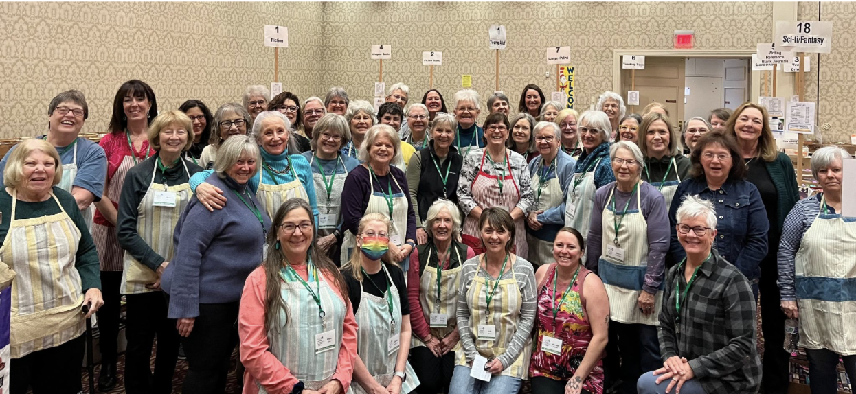 Group of women smiling at a conference or event, some wearing aprons, in a room with signs for different categories like fiction and sci-fi.
