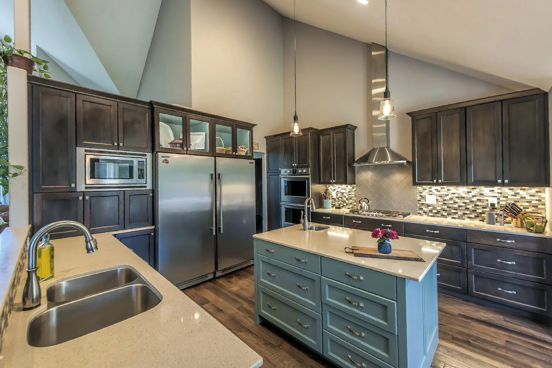 Modern kitchen with dark wood cabinets, stainless steel appliances, a central island with a light blue base, and a decorative tile backsplash. The kitchen features an open ceiling design with pendant lighting.