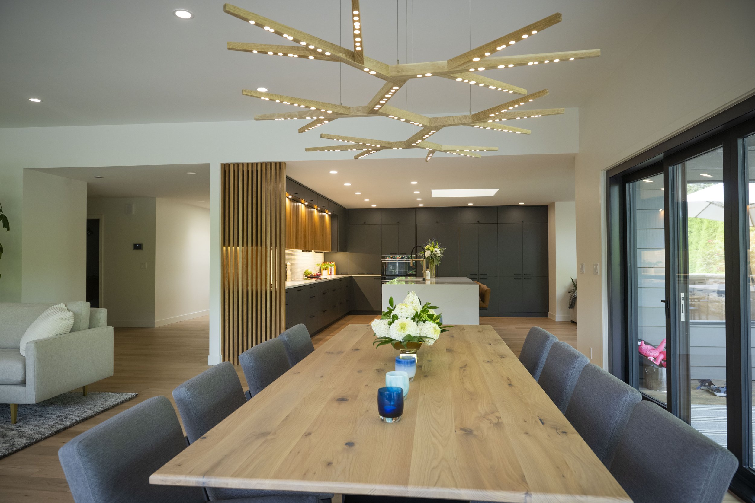 Modern open-concept kitchen and dining area with a large wooden table surrounded by gray upholstered chairs, a flower centerpiece, contemporary chandelier, black cabinetry, sliding glass doors, and a partial view of living space.