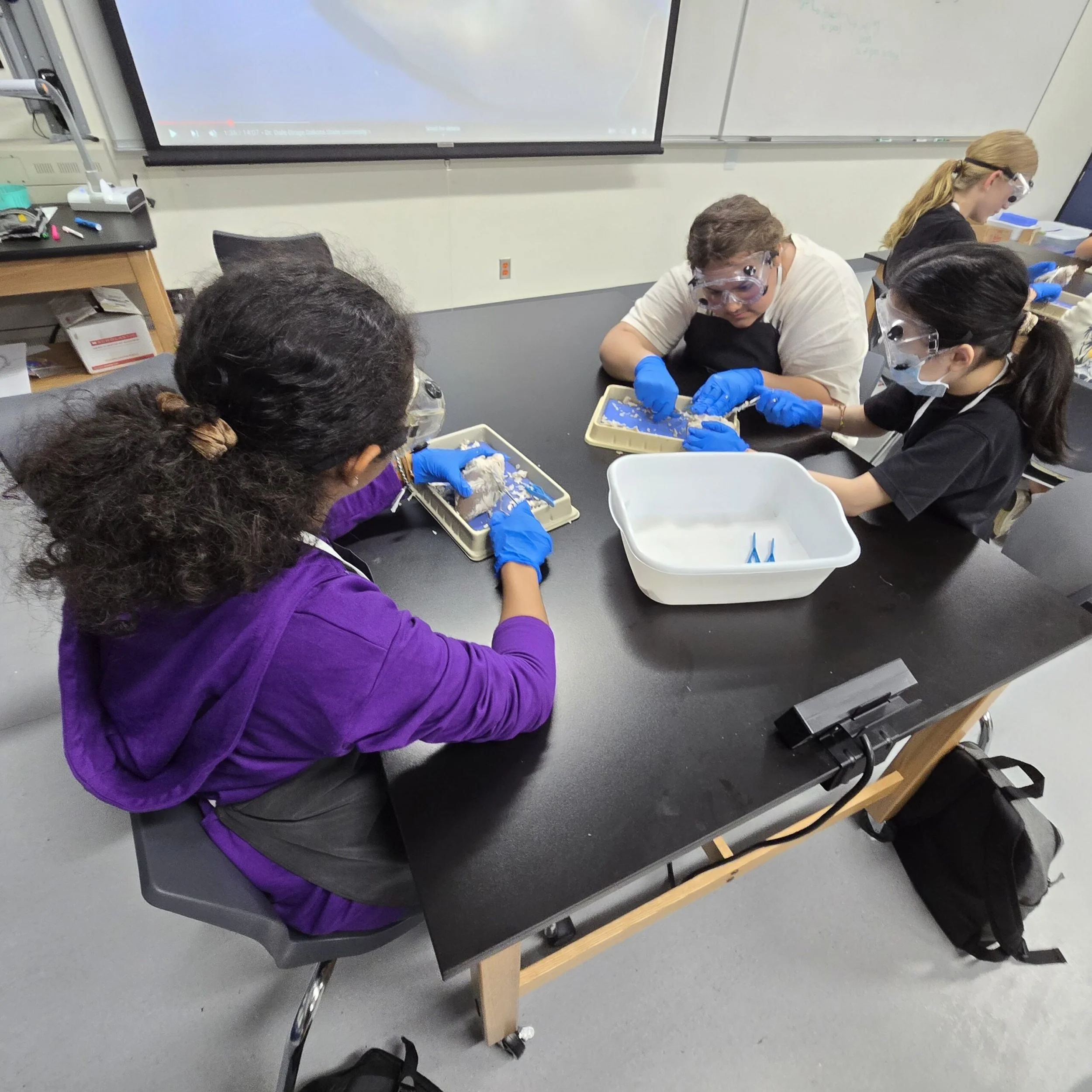 Students in lab coats and safety goggles working with scientific specimens at laboratory tables.
