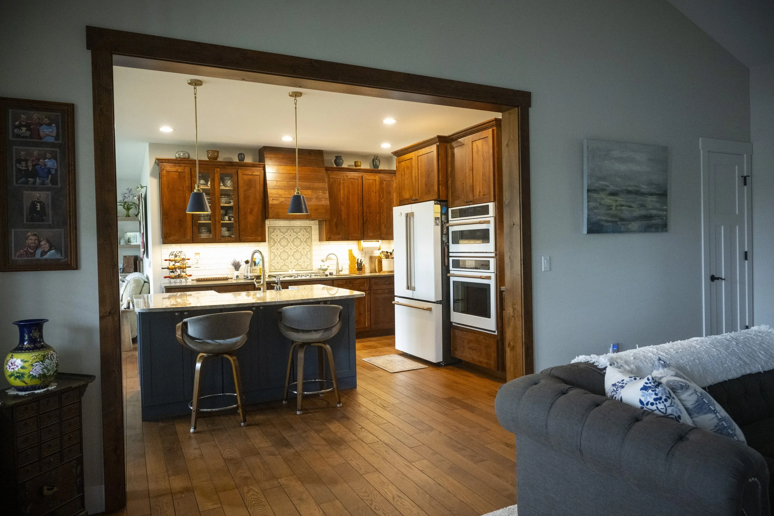 Open-concept kitchen with wooden cabinets, white appliances, and a kitchen island with bar stools, adjacent to a living room with a gray armchair and a wall hanging painting.