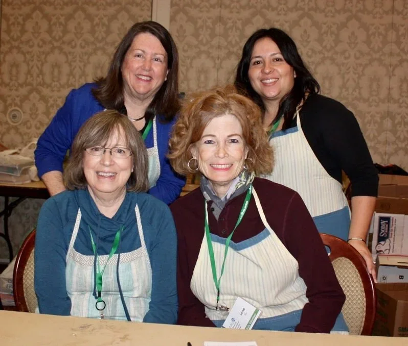 Four women, all wearing aprons with green lanyards, smiling and posing together in a room with patterned wallpaper, sitting and standing around a wooden table.