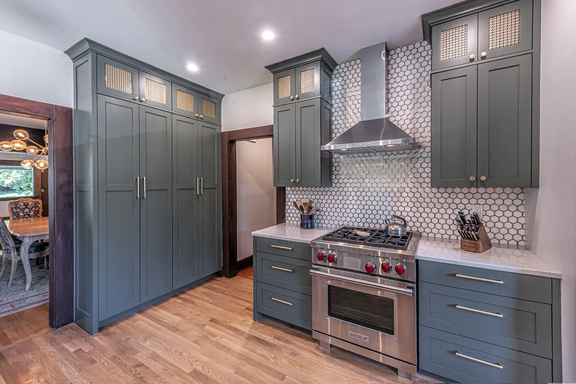 View of a modern kitchen with blue cabinets, a stainless steel stove, and a hexagonal tile backsplash. The kitchen has wood flooring and recessed lighting in the ceiling.