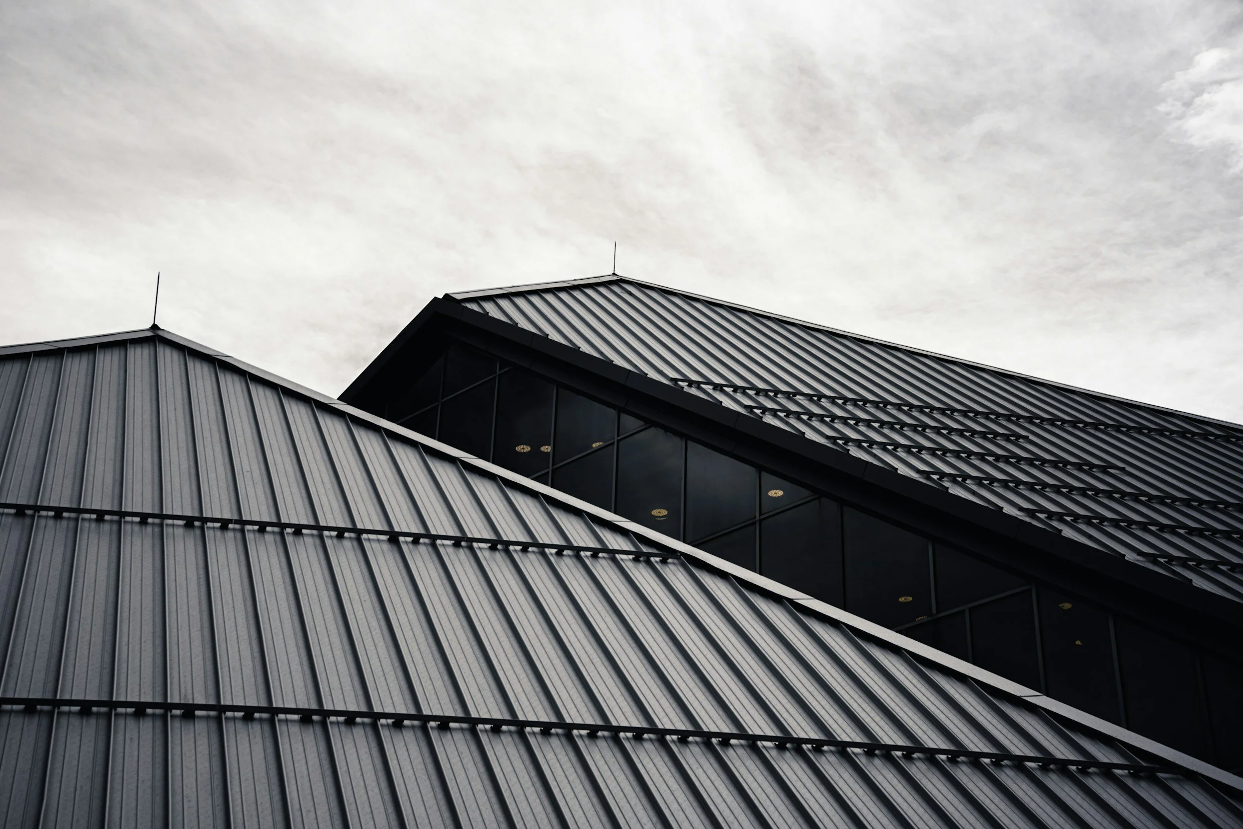 A modern building with metal roofing panels and large glass window sections, viewed from below against a cloudy sky.