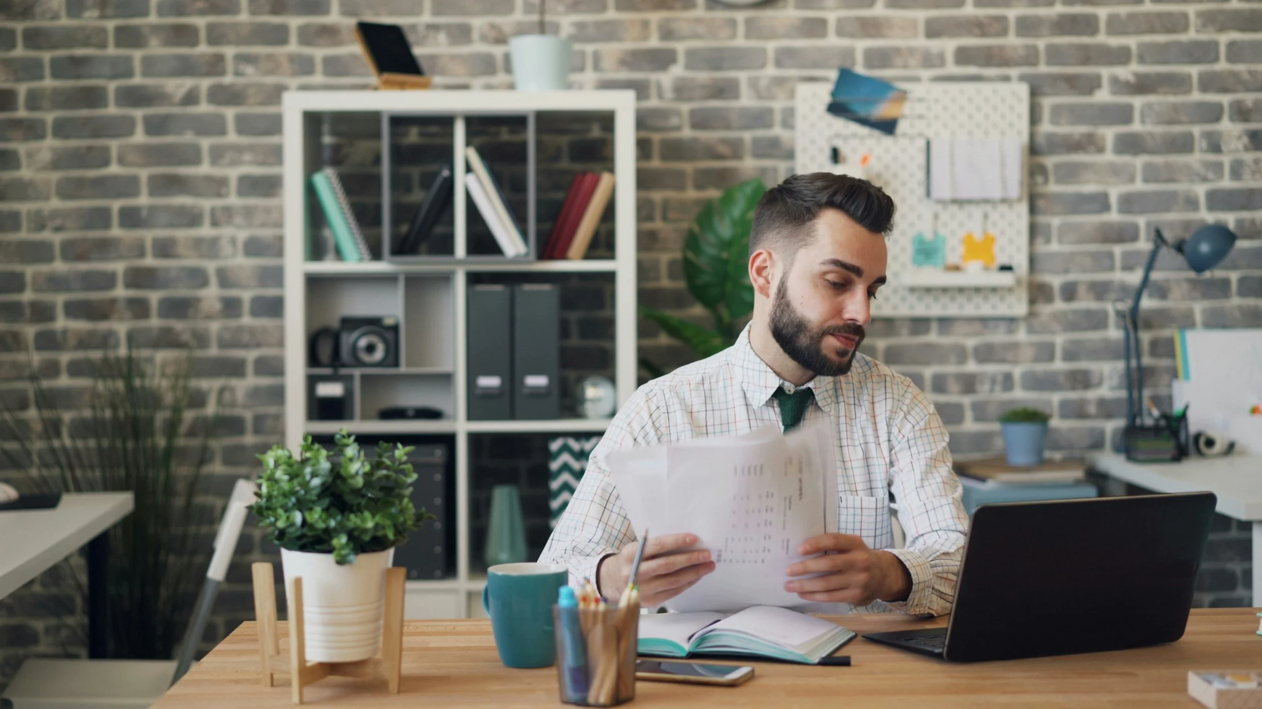 A man in a checkered shirt sits at a wooden desk with an open notebook, a laptop, a mug, and some stationery. Behind him is a bookshelf with books and a camera, and a whiteboard with notes. The room has a brick wall and looks like a modern office or home workspace.