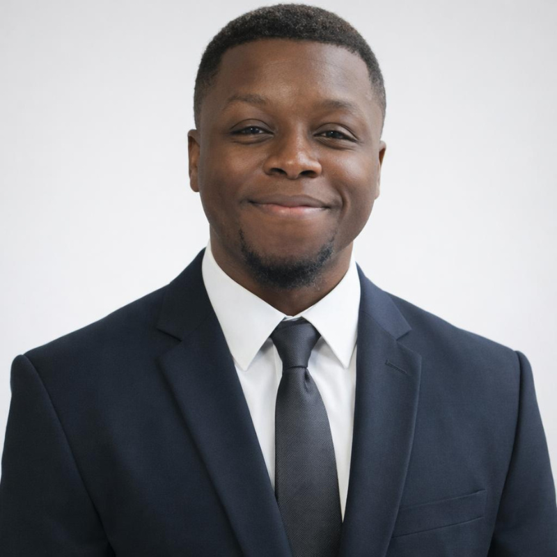 Portrait of a smiling young Black man in a business suit, white shirt, and dark tie against a plain white background.