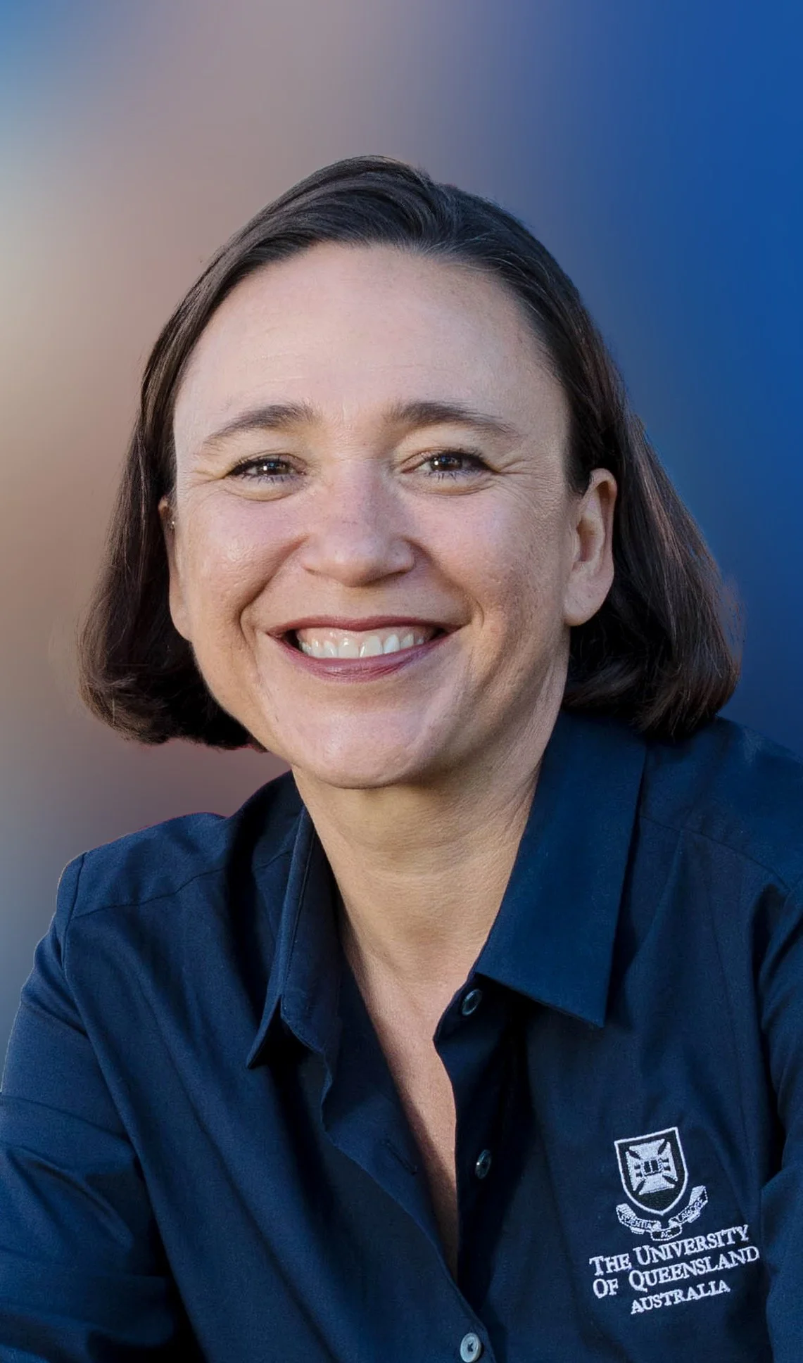 A woman smiling in a dark blue shirt with the logo of The University of Queensland, Australia, against a blurred outdoor background.