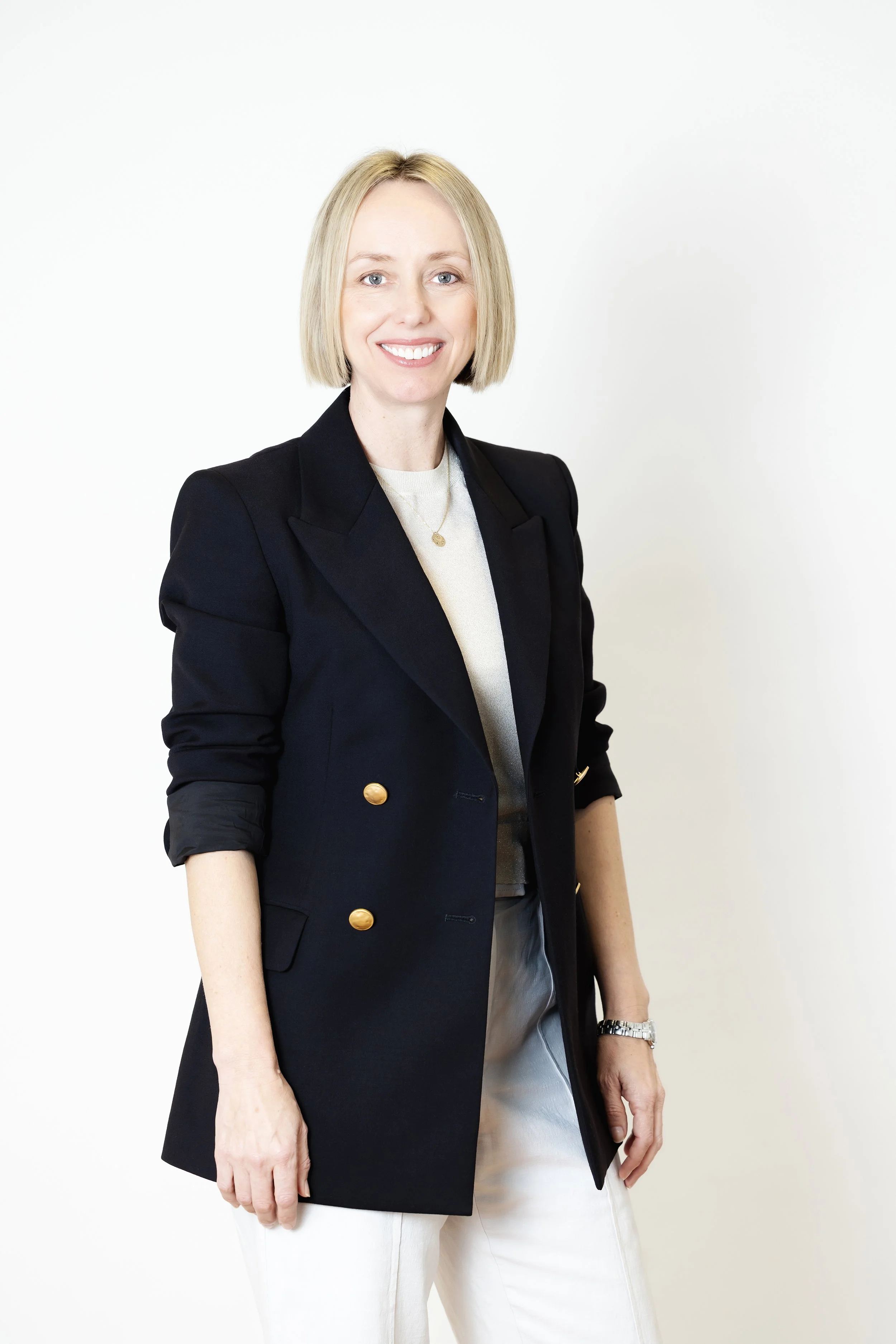A woman with blonde hair, smiling, wearing a black blazer with gold buttons, a white top, and white pants, standing against a plain white background.