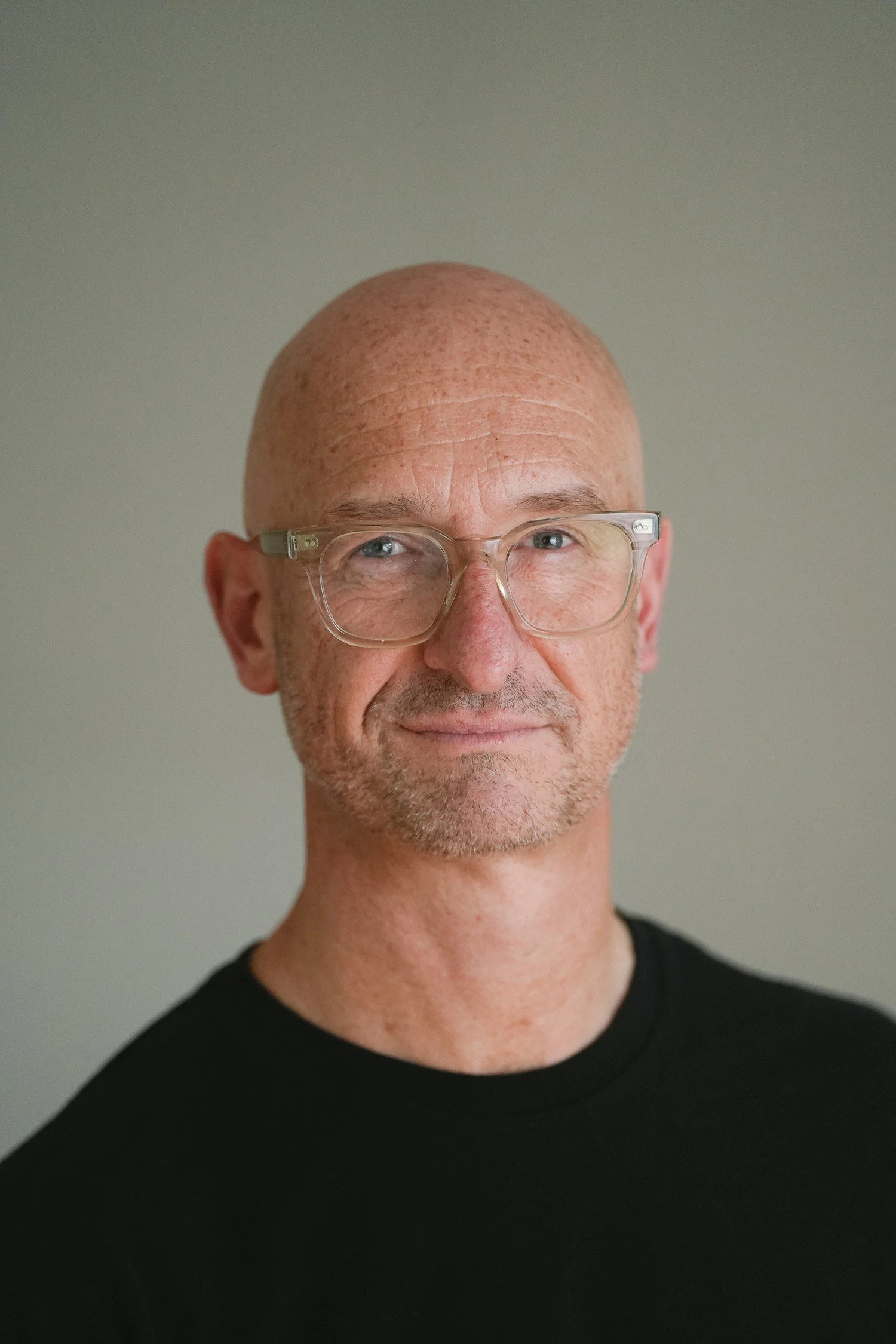 Portrait of a bald man with clear glasses, light skin, faint beard, wearing a black shirt, smiling softly against a plain background.