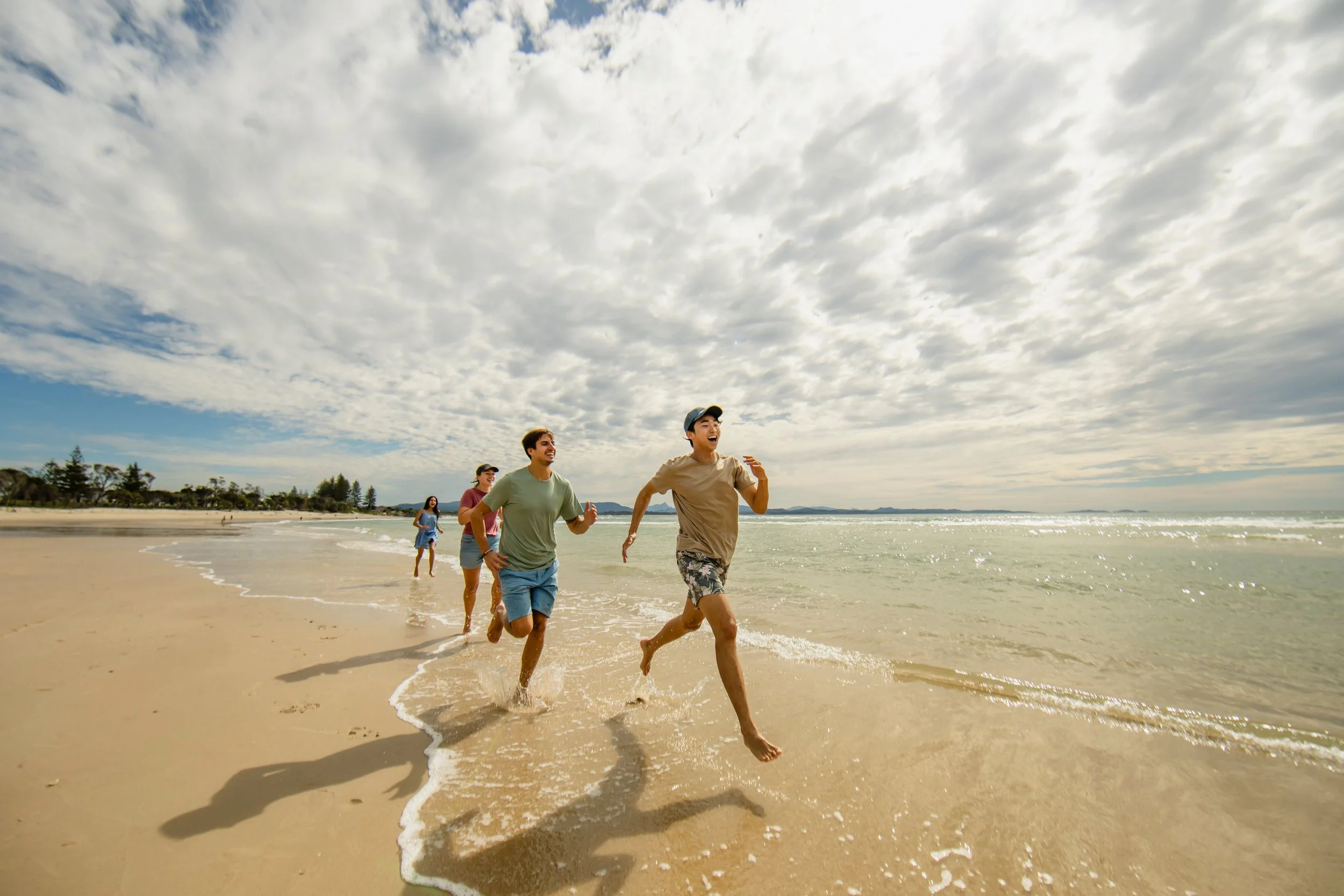 A group of five friends running along the shoreline at the beach during the daytime, with cloudy skies overhead.