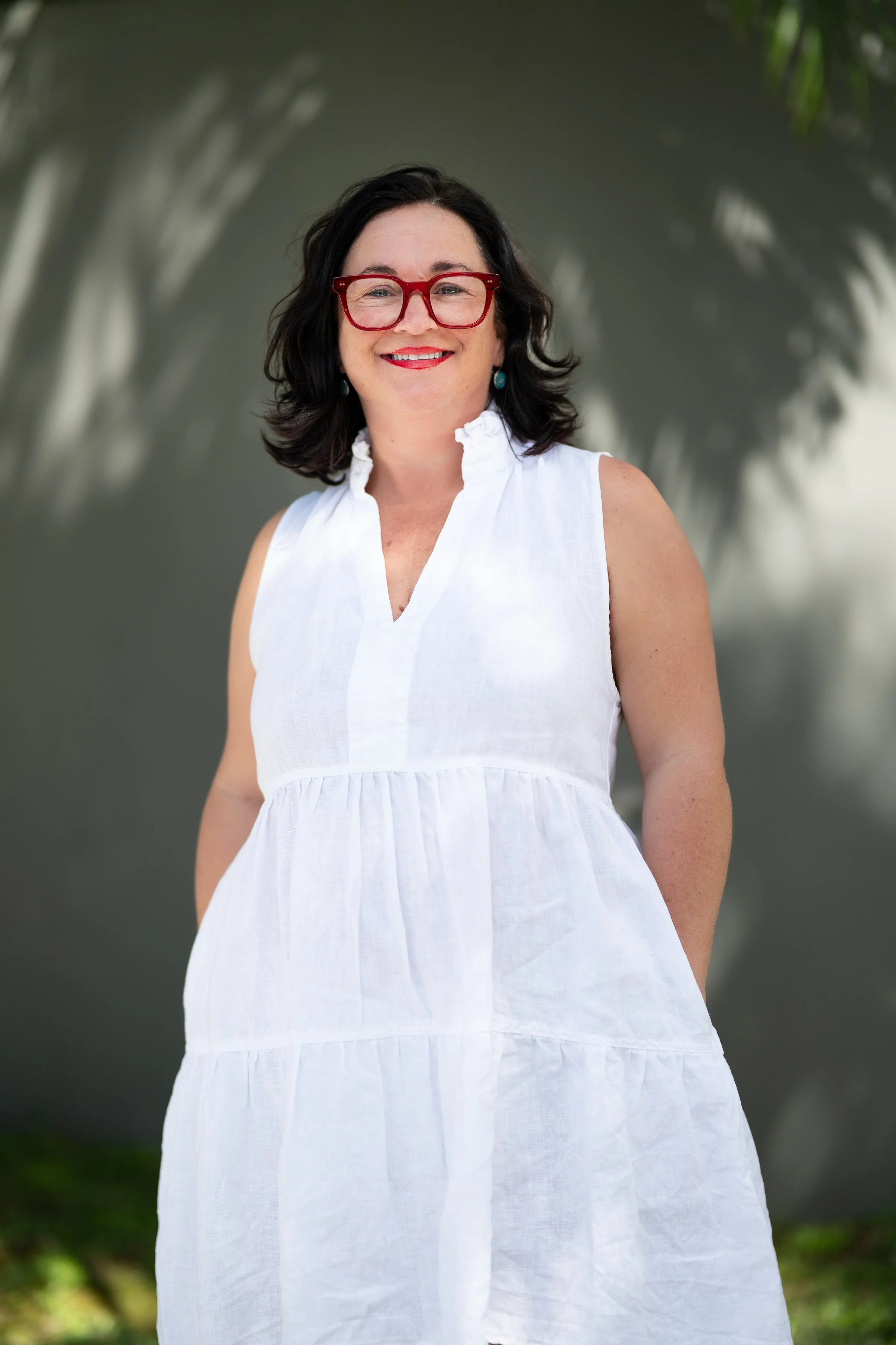 A woman with dark hair wearing red glasses and a white sleeveless dress, smiling outdoors with shadowed foliage in the background.
