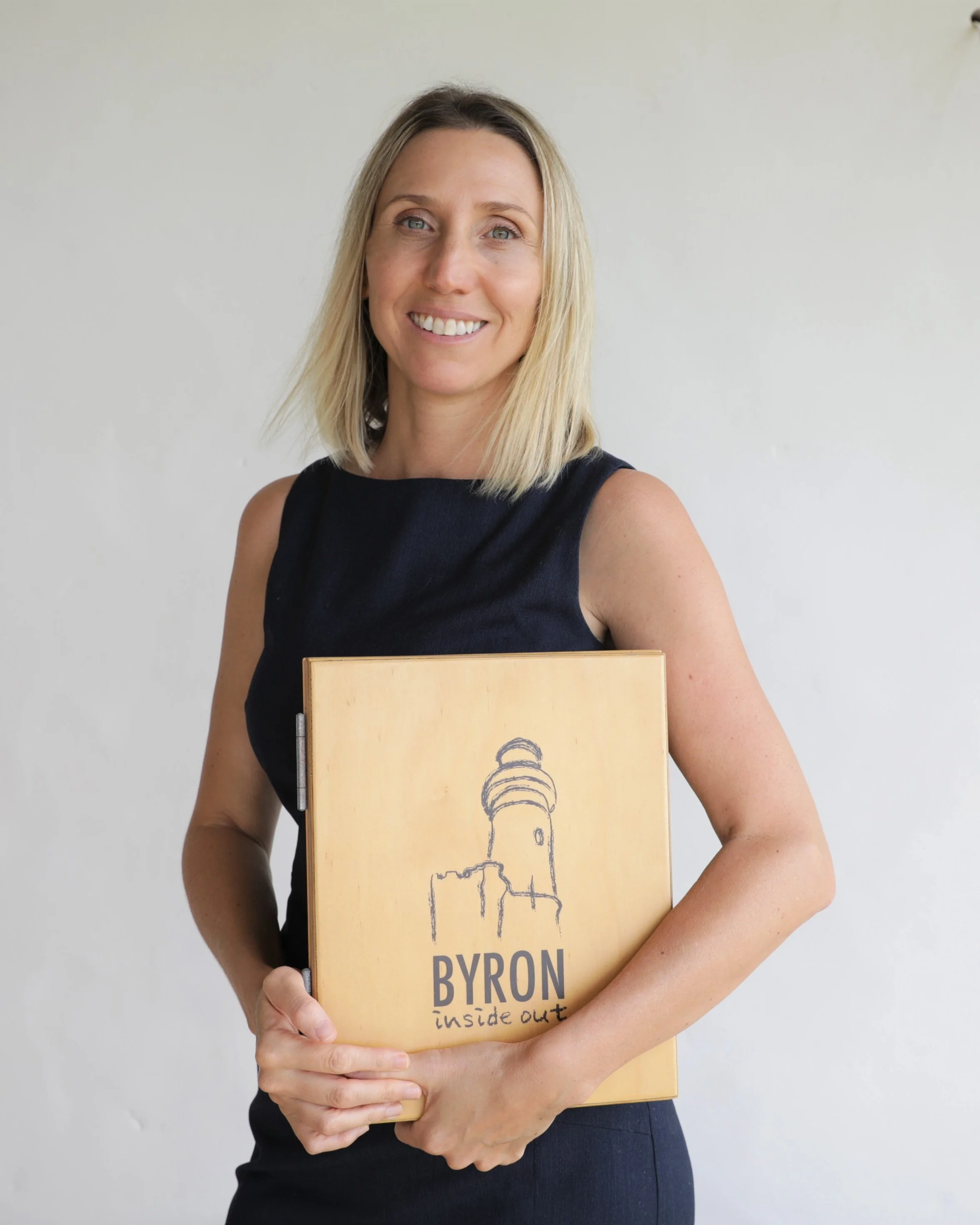 A woman with blonde hair and a black sleeveless top holding a folder with a sketch of a lighthouse and the words 'BYRON inside out' written on it.