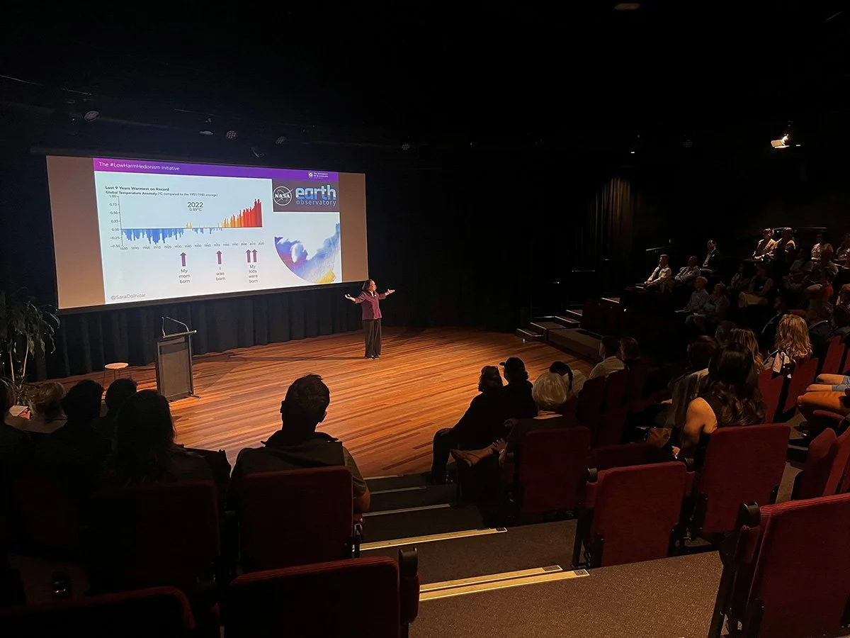 A woman giving a presentation on climate change at a conference, with a slide showing global temperature anomalies and the NASA Earth Observatory logo, in a dark auditorium with an audience seated in red chairs.