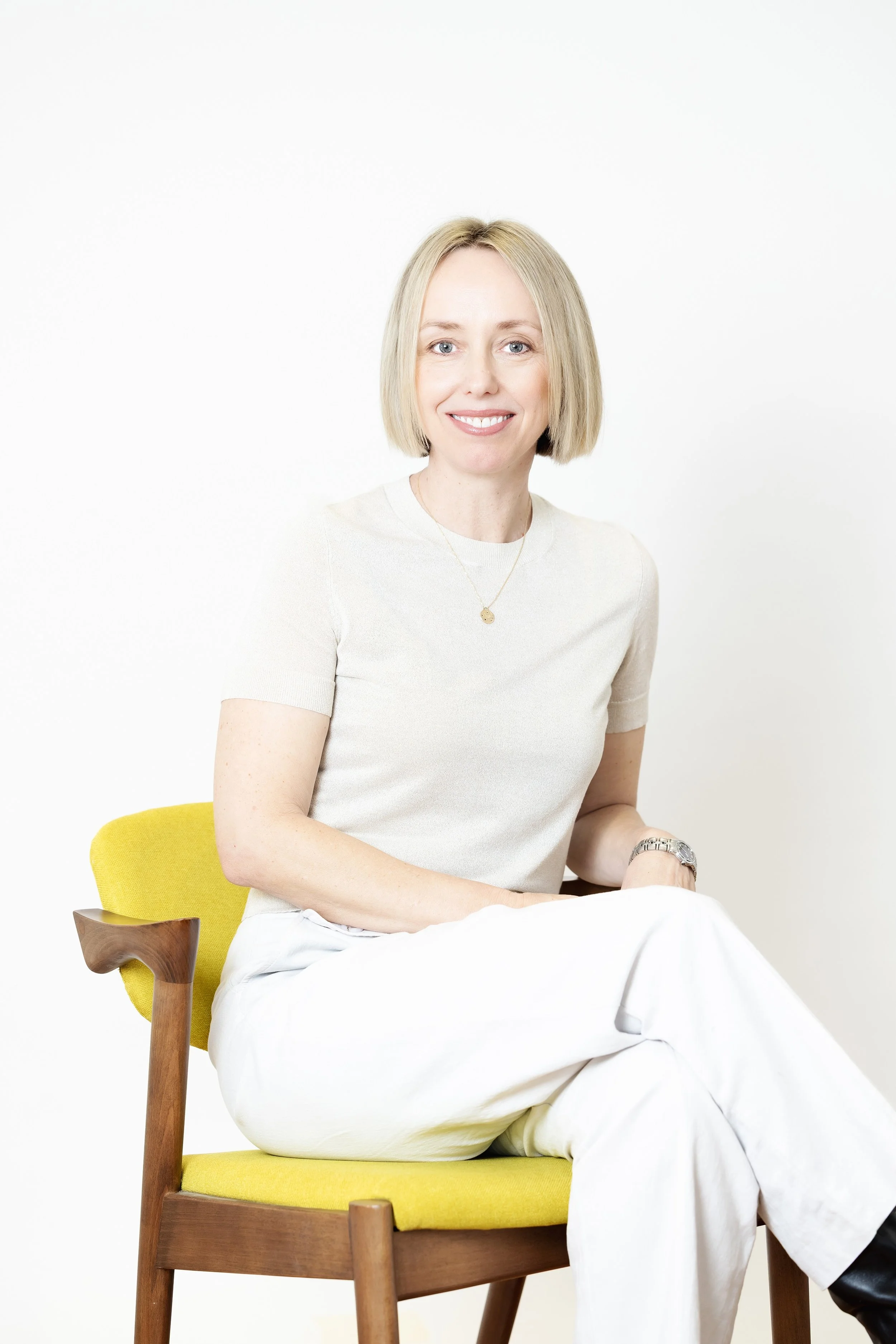 A woman with blonde hair sitting on a yellow chair with wooden armrests, wearing a white short-sleeve top, white pants, a silver watch, and a necklace, smiling against a plain white background.