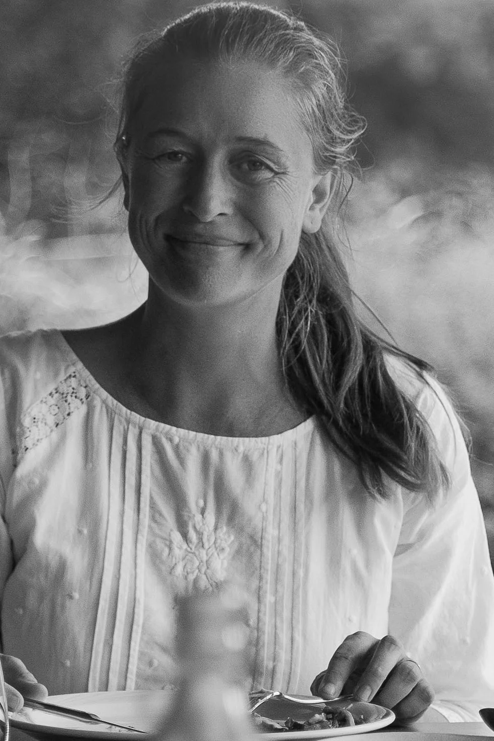 A smiling woman with long hair in a casual top, sitting at a table with a plate of food, outdoors with blurred background.