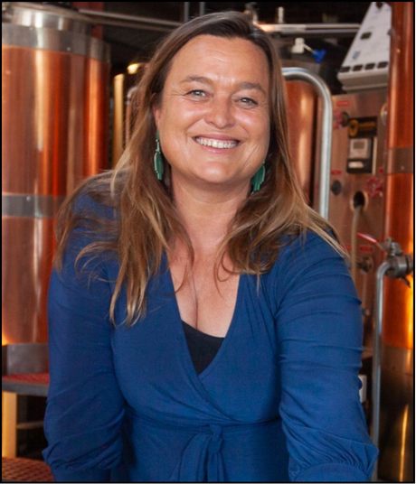 A woman with long blonde hair smiling, wearing a blue blazer and teal earrings, standing in front of copper brewing equipment.