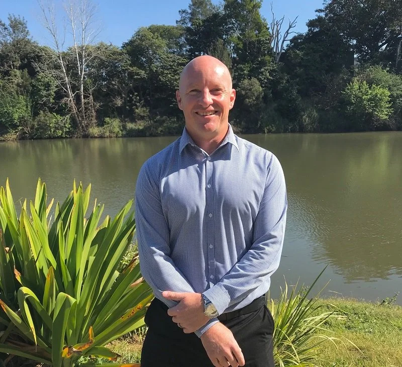 A bald man wearing a blue checkered dress shirt and black pants standing near a lake with green plants and trees in the background.
